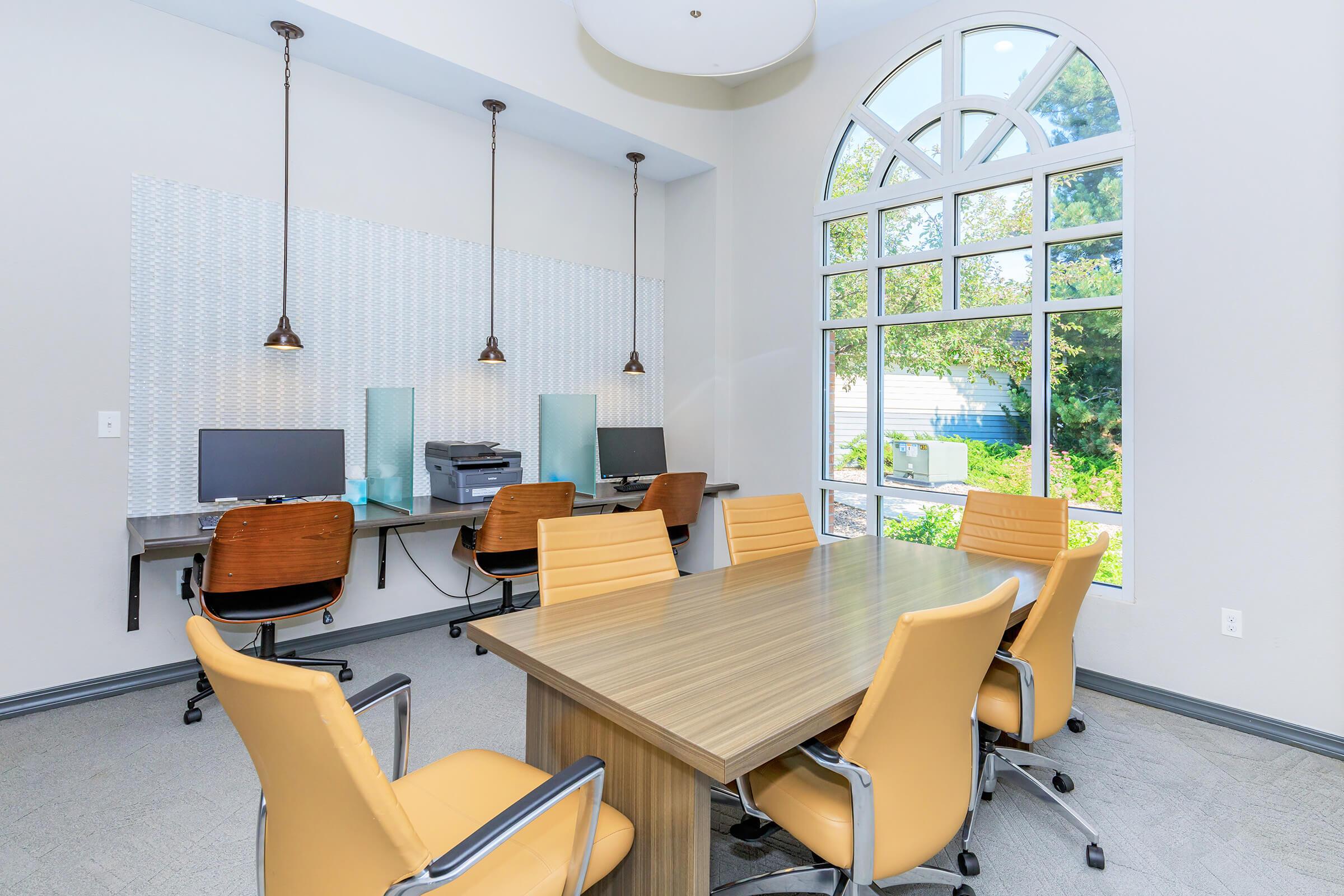 A modern conference room featuring a large wooden table surrounded by yellow ergonomic chairs. Two workstations with computers and a printer are positioned against the wall, illuminated by pendant lights. Large arched windows allow natural light to fill the space, with greenery visible outside.