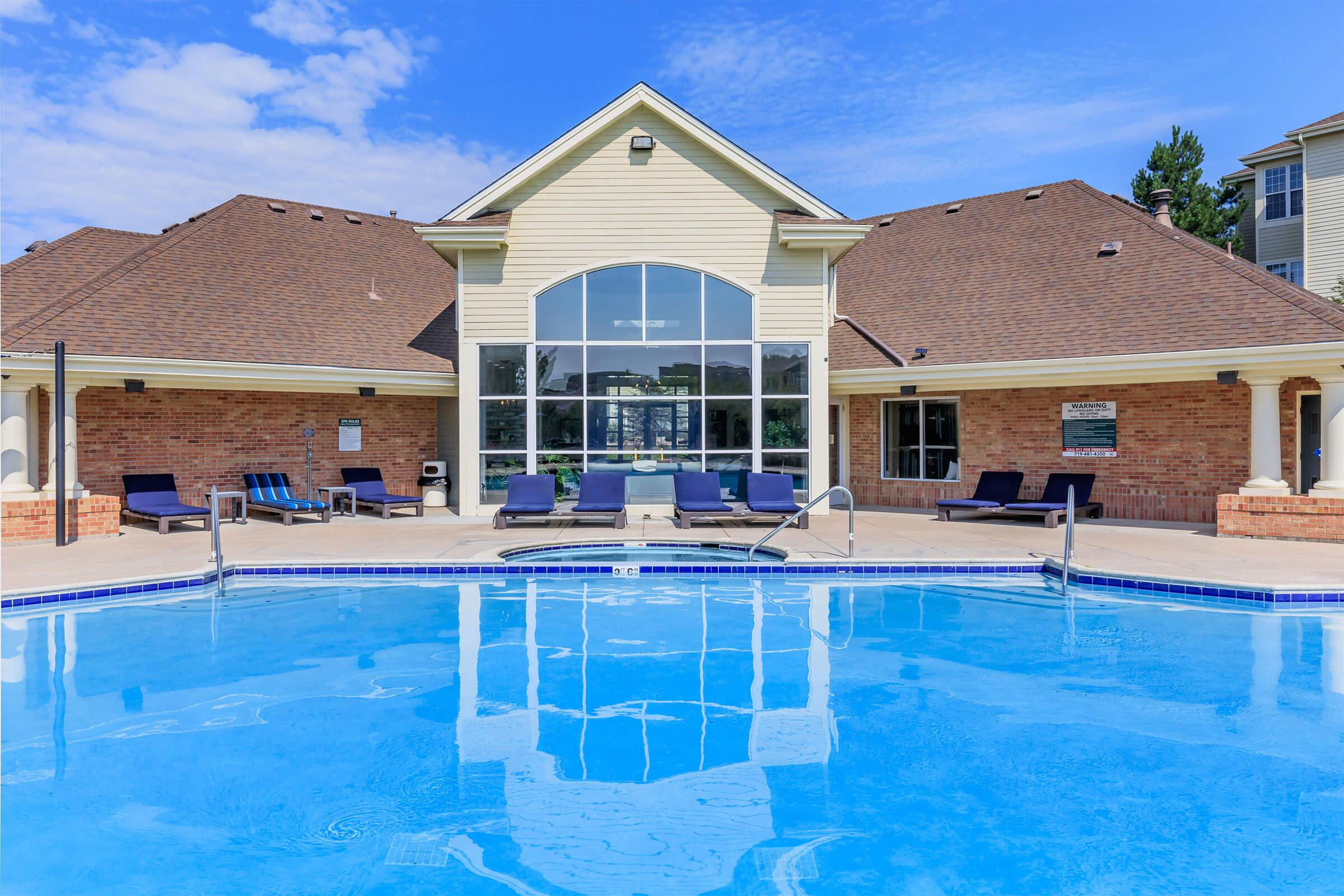 A clear blue swimming pool in the foreground, reflecting the sky. In the background, a modern building with a large arched window, surrounded by lounge chairs. The scene features well-maintained landscaping, with a sunny sky and a few scattered clouds, creating a relaxing outdoor atmosphere.