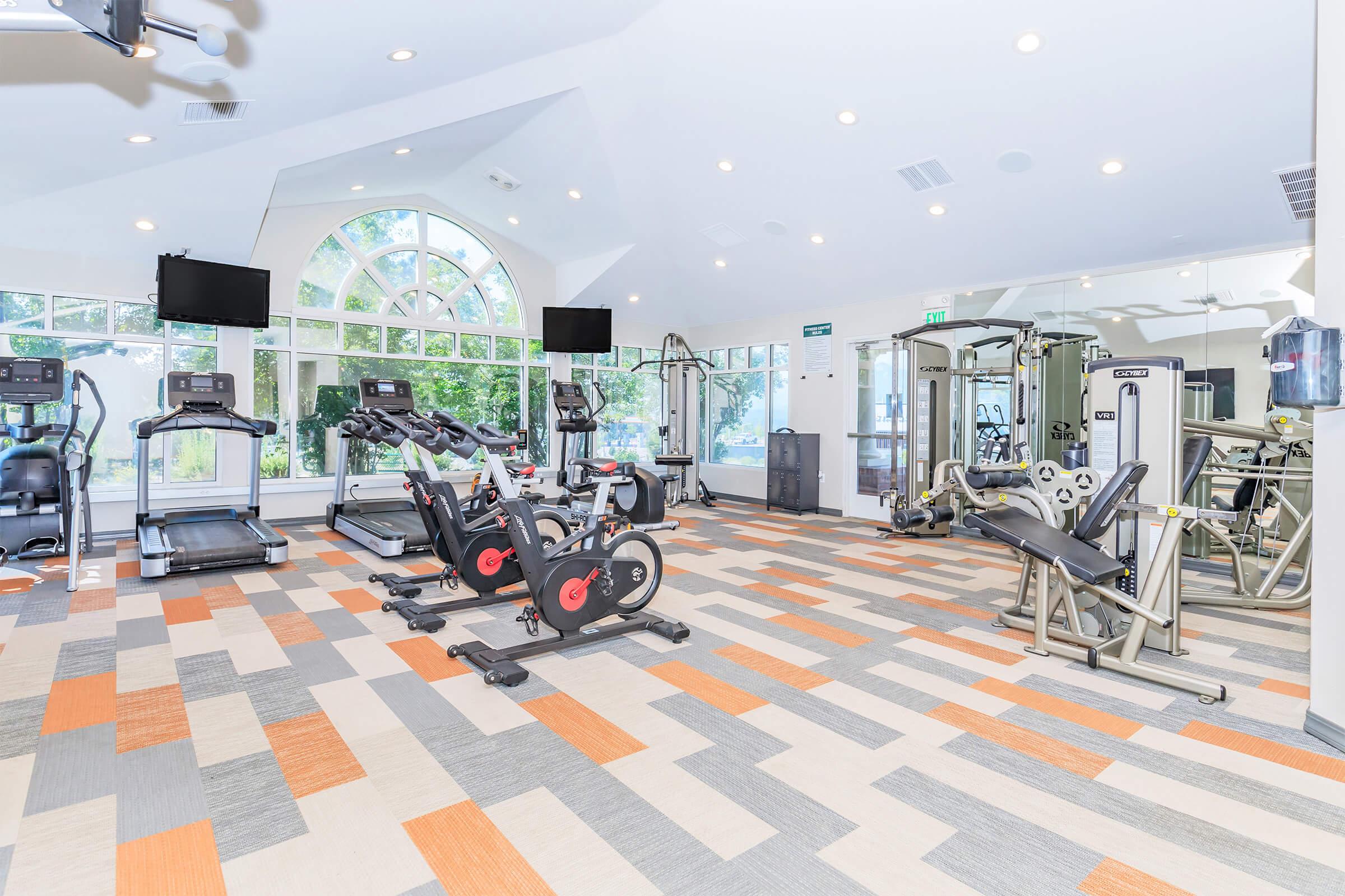 A modern gym interior featuring various exercise machines, including treadmills, stationary bikes, and strength training equipment. The space has bright lighting, large windows letting in natural light, and a colorful, patterned floor design.