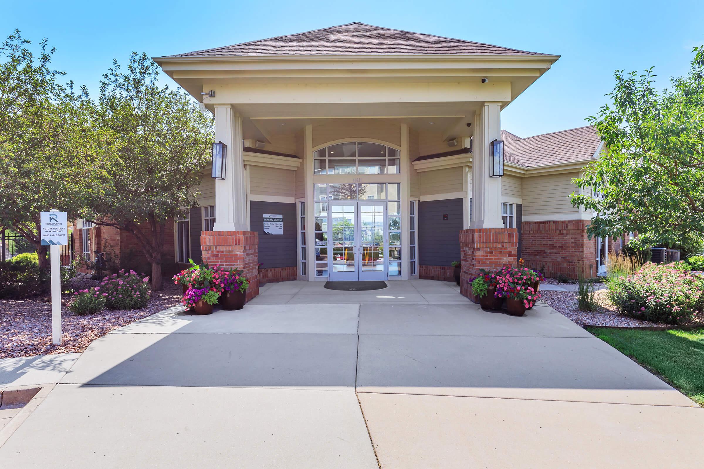 A welcoming entrance to a building featuring large glass doors, surrounded by well-maintained flower beds. The architecture includes a sloped roof and decorative elements, suggesting an inviting atmosphere. Sunlight brightens the scene, highlighting a clean concrete pathway leading to the entrance.