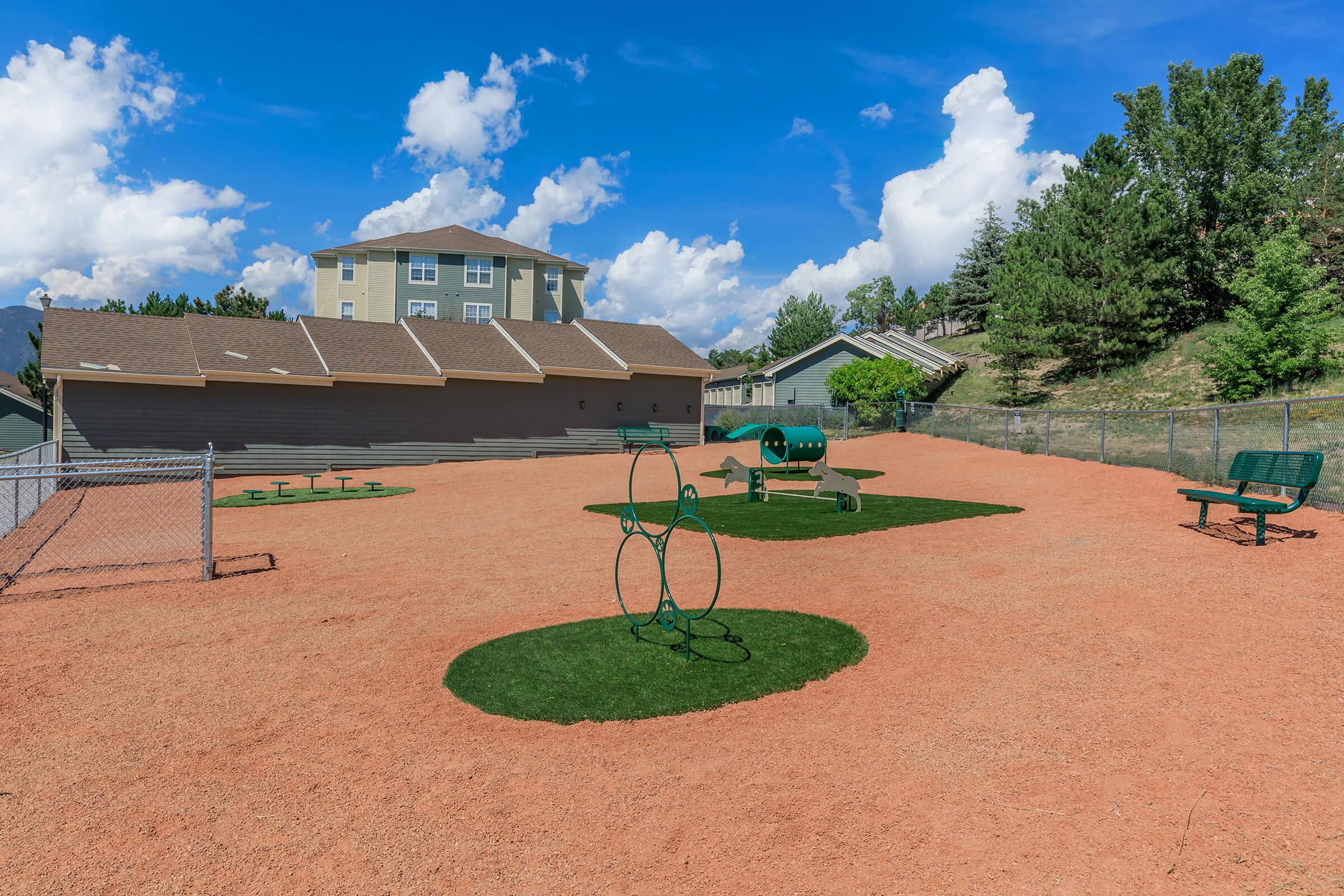 A bright park with a sandy surface, featuring various green play structures including a looped climbing frame and a round spinning apparatus. In the background, there are benches, tables, and several trees, under a clear blue sky with fluffy clouds. A building can be seen in the distance.