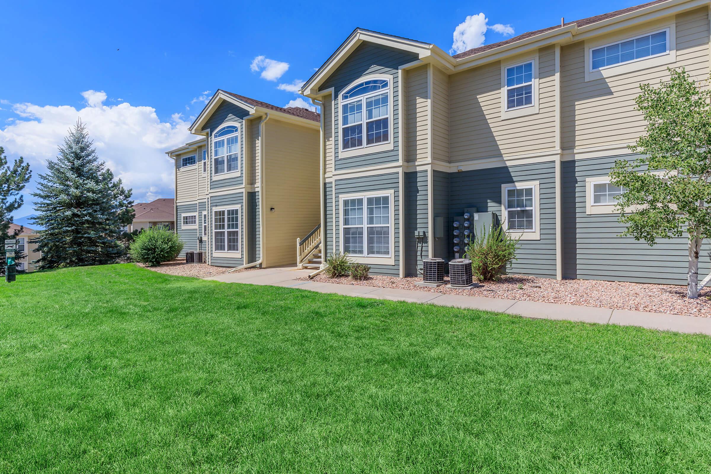 A well-maintained multi-unit residential building with light yellow and brown siding. The foreground features a manicured green lawn, and the background includes a clear blue sky with scattered clouds. There are shrubs and trees around the building, contributing to a pleasant residential environment.