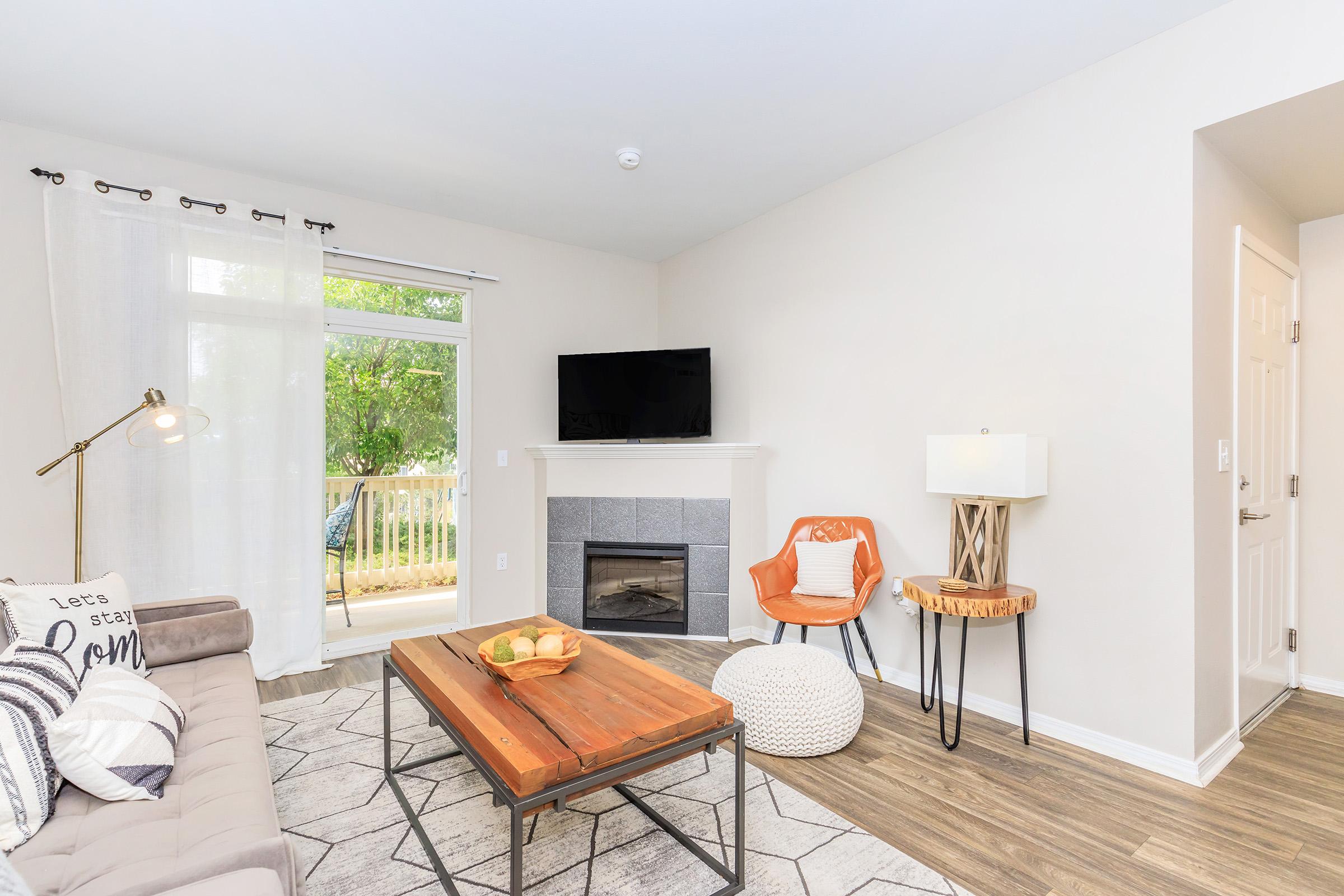 A cozy living room featuring a gray sofa, a wooden coffee table, and a decorative pouf. There is an orange accent chair beside a lamp and a small table. A fireplace is integrated into the wall, and a flat-screen TV is mounted above it. Natural light floods in from a nearby window overlooking a balcony.