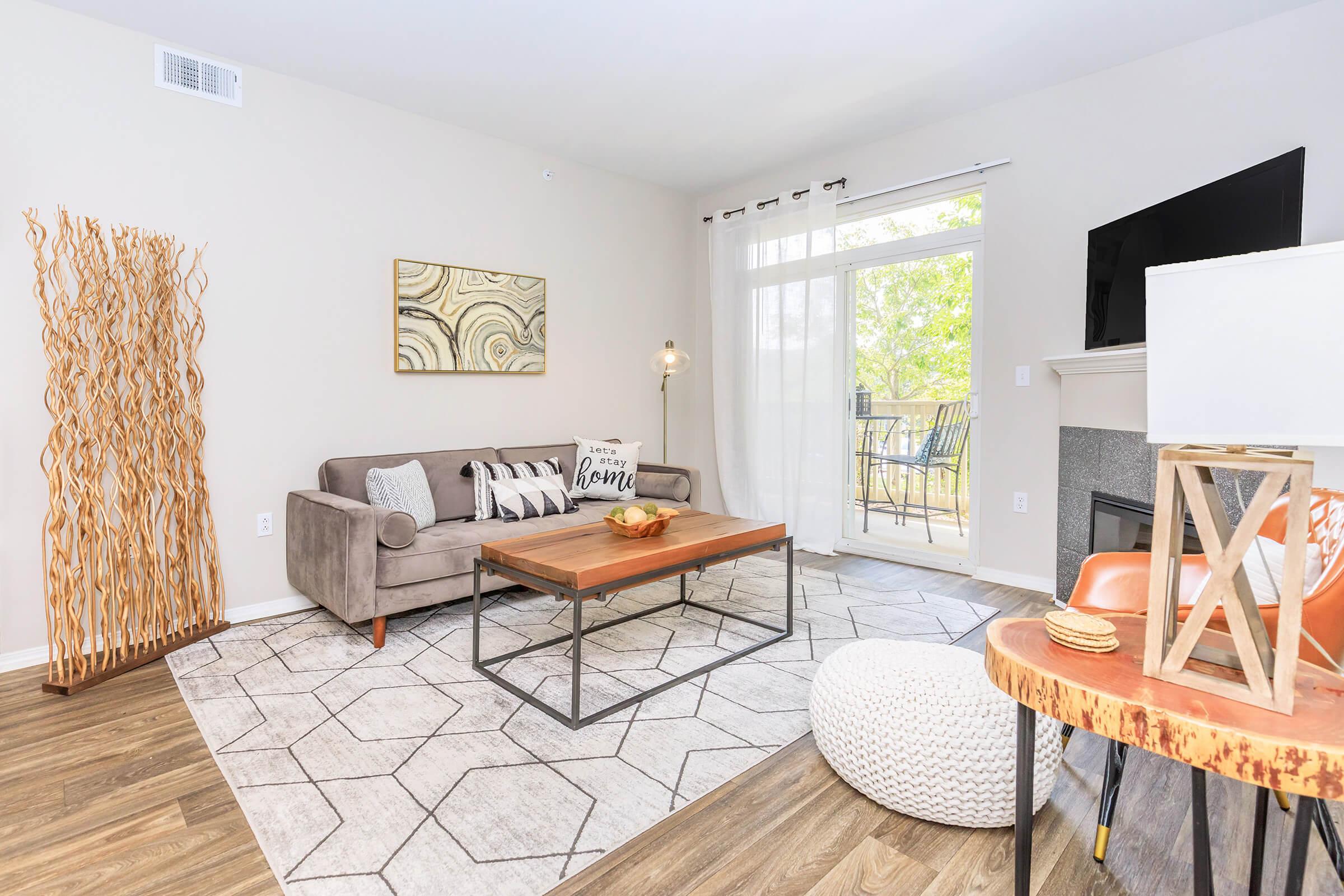 A cozy living room featuring a gray sofa with decorative pillows, a wooden coffee table, and a light-colored rug with geometric patterns. There's a large window with sheer curtains letting in natural light, a small round pouf, and a fireplace with a decorative lantern nearby.