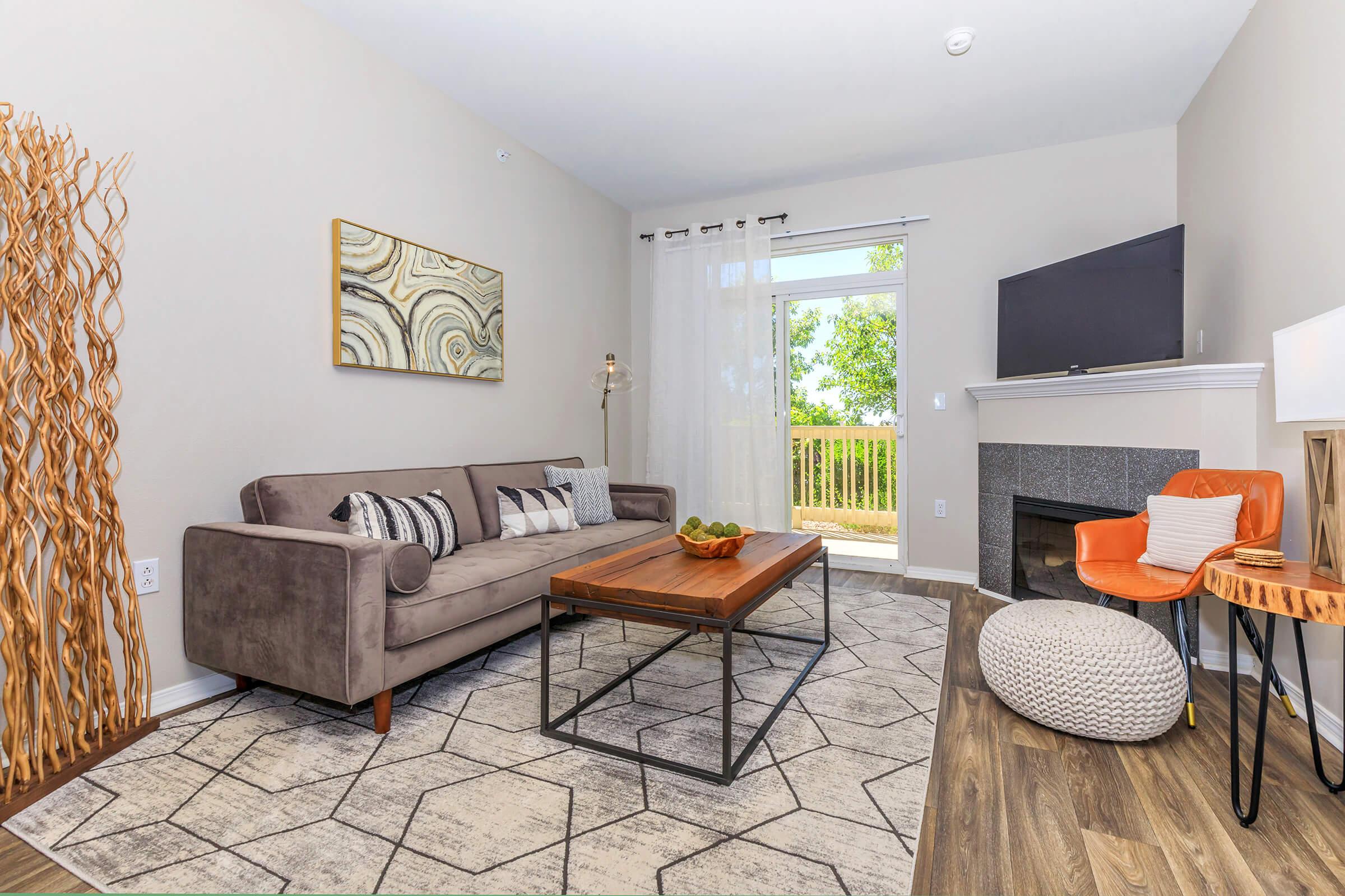 A modern living room featuring a stylish gray sofa with decorative pillows, a wooden coffee table, and a cozy accent chair. There’s a textured area rug on the floor, a flat-screen TV mounted on the wall, and large windows allowing natural light, with a view of greenery outside.