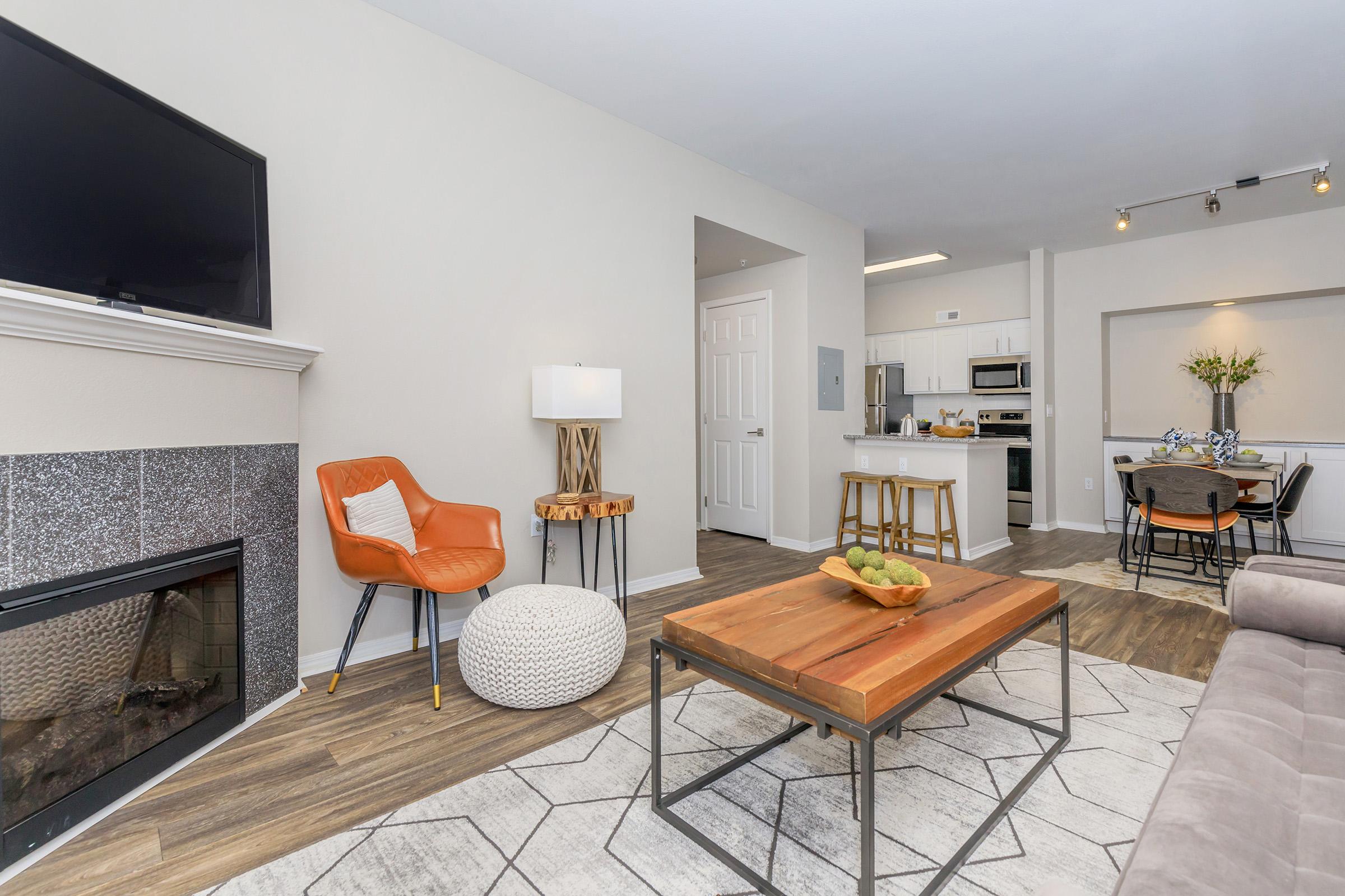 Living room featuring a modern design with a flat-screen TV mounted above a fireplace, an orange accent chair, a round white pouf, and a wooden coffee table. A dining area can be seen in the background, with a table and chairs. Light walls and wood flooring enhance the contemporary aesthetic.