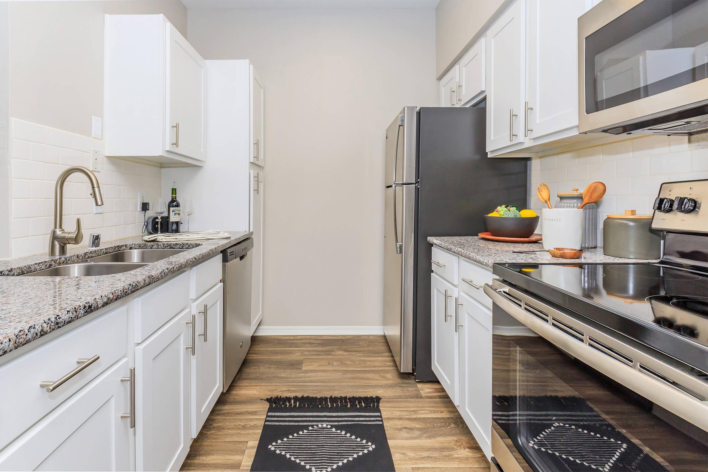 A modern kitchen featuring white cabinetry, stainless steel appliances, and a granite countertop. The space includes a sink, dishwasher, and a stove with an oven. A black and white patterned runner is placed on the floor, and a bowl of fruit is on the counter, enhancing the inviting atmosphere.