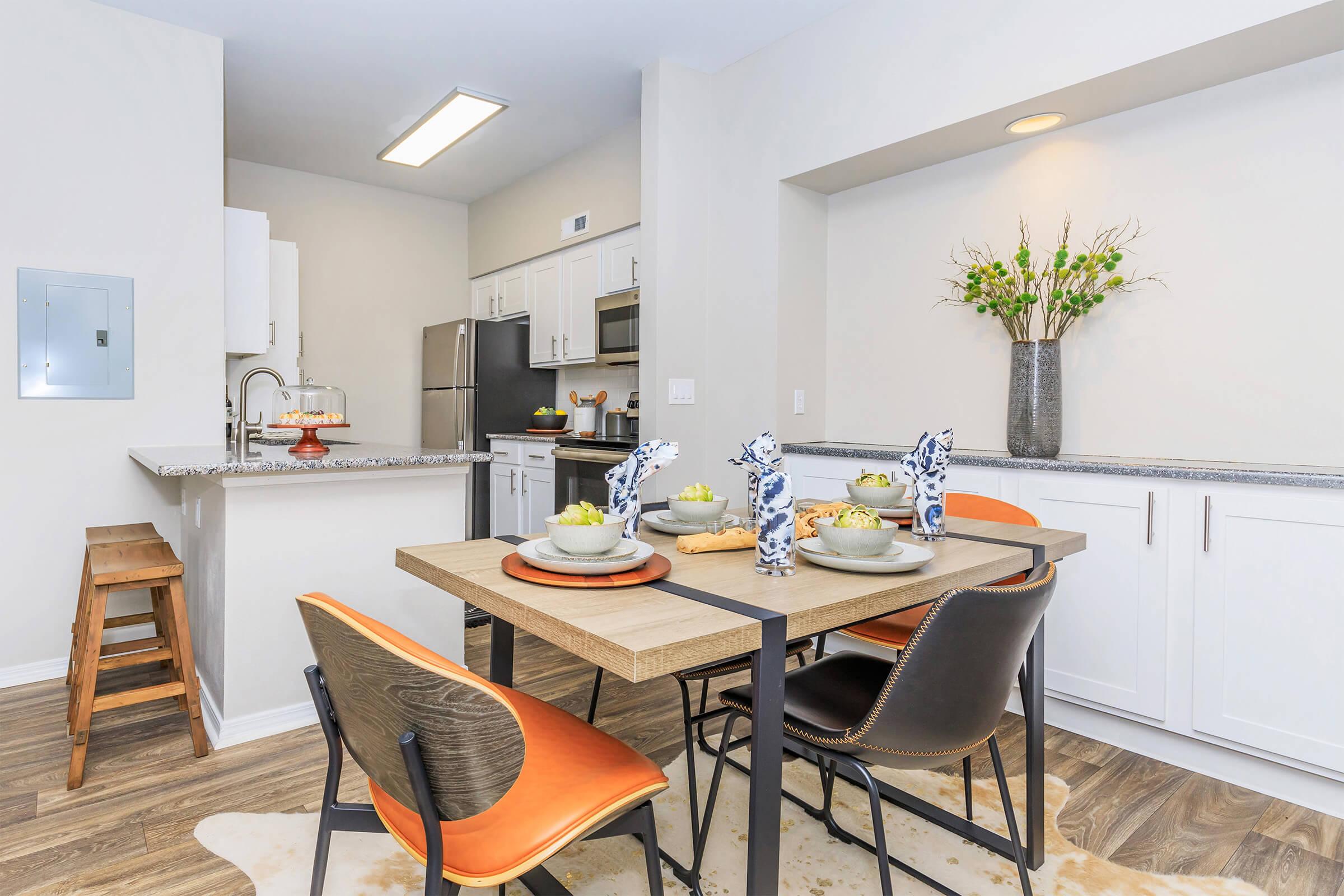 A modern kitchen and dining area featuring a table set for four with decorative plates and bowls, surrounded by stylish black and orange chairs. A small bar stool is in the corner, and the kitchen includes white cabinets, stainless steel appliances, and decorative plants on the countertop.