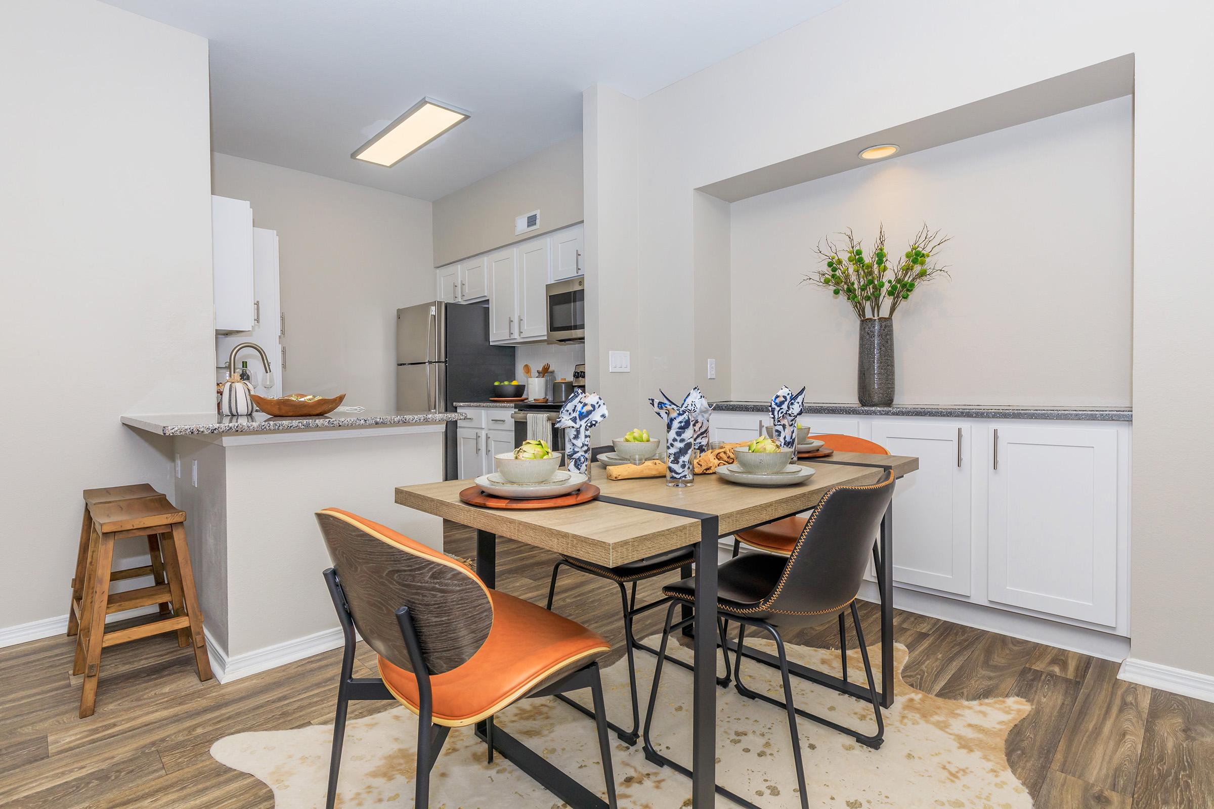 A modern kitchen and dining area featuring a wooden table set for a meal with plates, glasses, and decorative items. The room has light-colored walls, sleek cabinetry, and stainless steel appliances. A stylish bar stool is visible, along with a decorative arrangement of plants. Natural lighting enhances the open, inviting space.