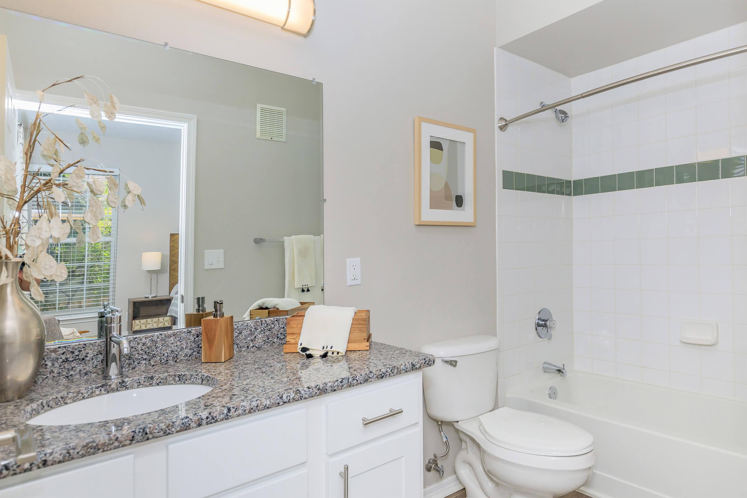 A modern bathroom featuring a white bathtub with a shower, a granite countertop with a sink, a decorative vase with dried flowers, and a framed art piece on the wall. Natural light comes through a window, and there are towels neatly arranged nearby.