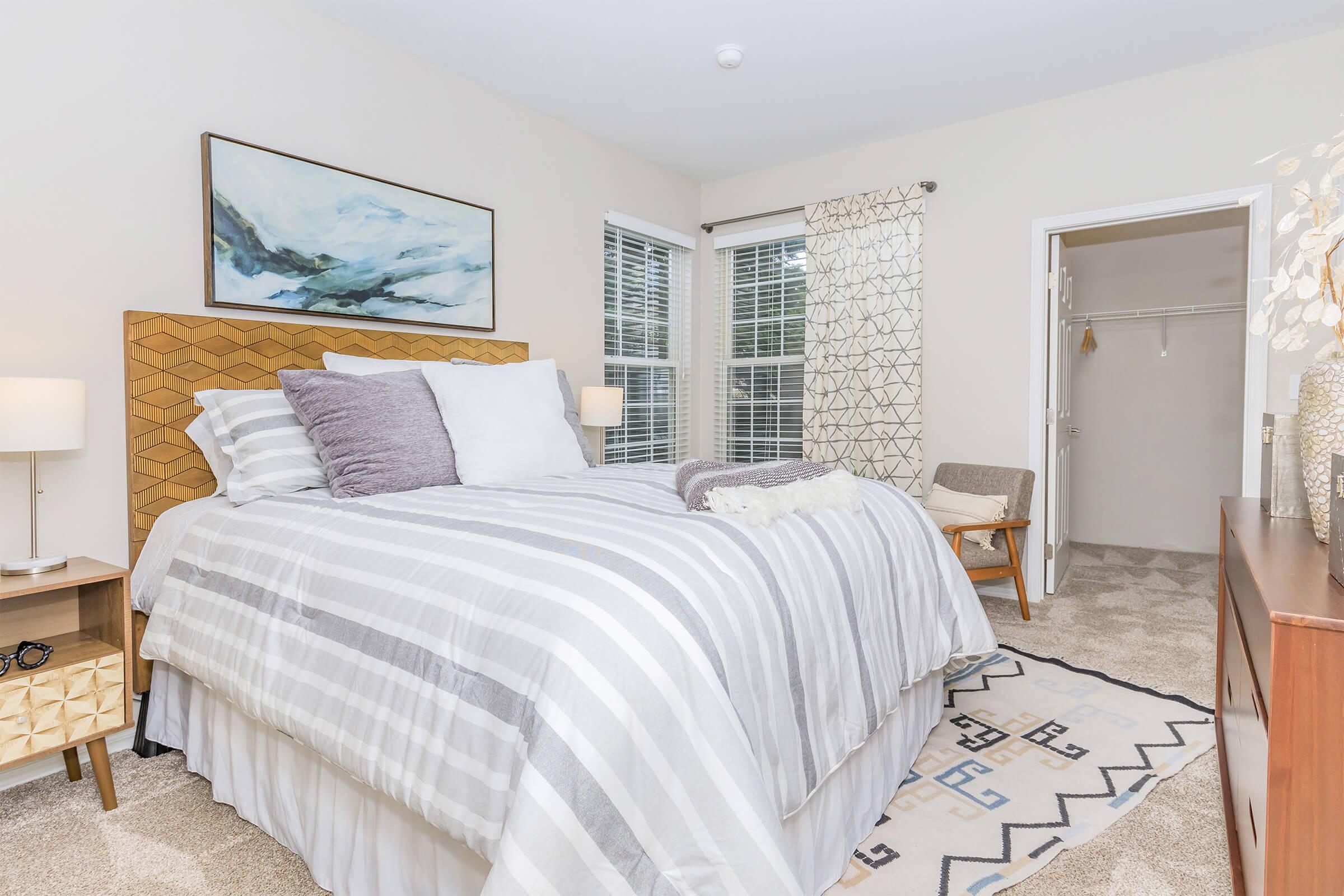 Spacious bedroom featuring a large bed with gray and white striped bedding, two bedside lamps, and a wooden nightstand. A framed landscape artwork hangs above the bed. Natural light streams in through windows adorned with sheer curtains. A closet is visible in the background, and a patterned rug adds warmth to the space.