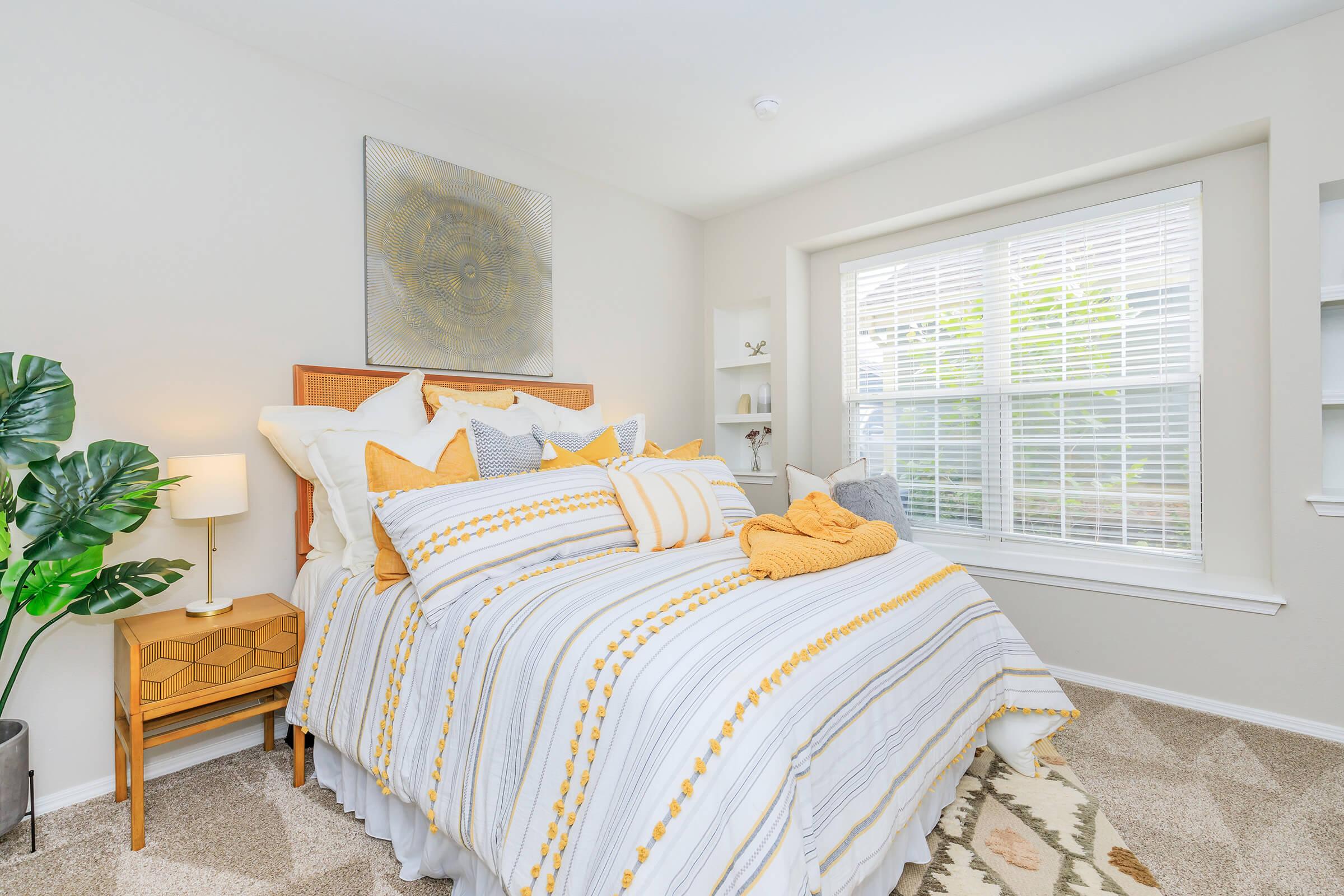 A cozy bedroom featuring a neatly made bed with yellow and white striped bedding and decorative pillows. There's a small bedside table with a lamp, a potted plant, and a window with white blinds letting in natural light. The walls are painted in soft neutral tones, creating a warm and inviting atmosphere.
