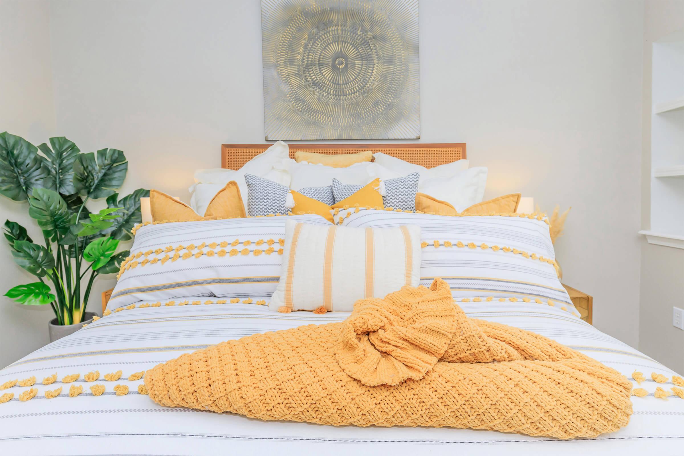 A cozy bed adorned with an array of yellow and white pillows and a knitted throw blanket. The bedding features striped patterns and decorative pom-poms. A green potted plant adds a touch of greenery, and a piece of wall art with a circular design hangs above the bed, creating a warm and inviting atmosphere.