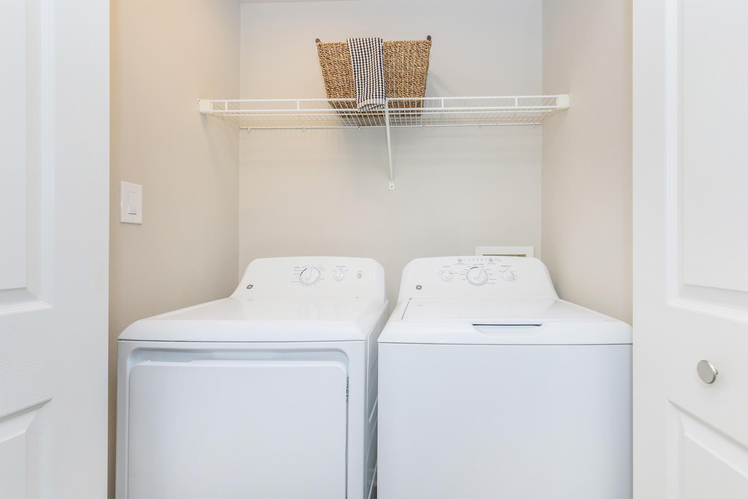 A compact laundry area featuring a white washing machine and dryer side by side. Above them is a wire shelf holding a woven basket. The walls are light-colored, and the space is organized, with white doors partially visible on the right side.