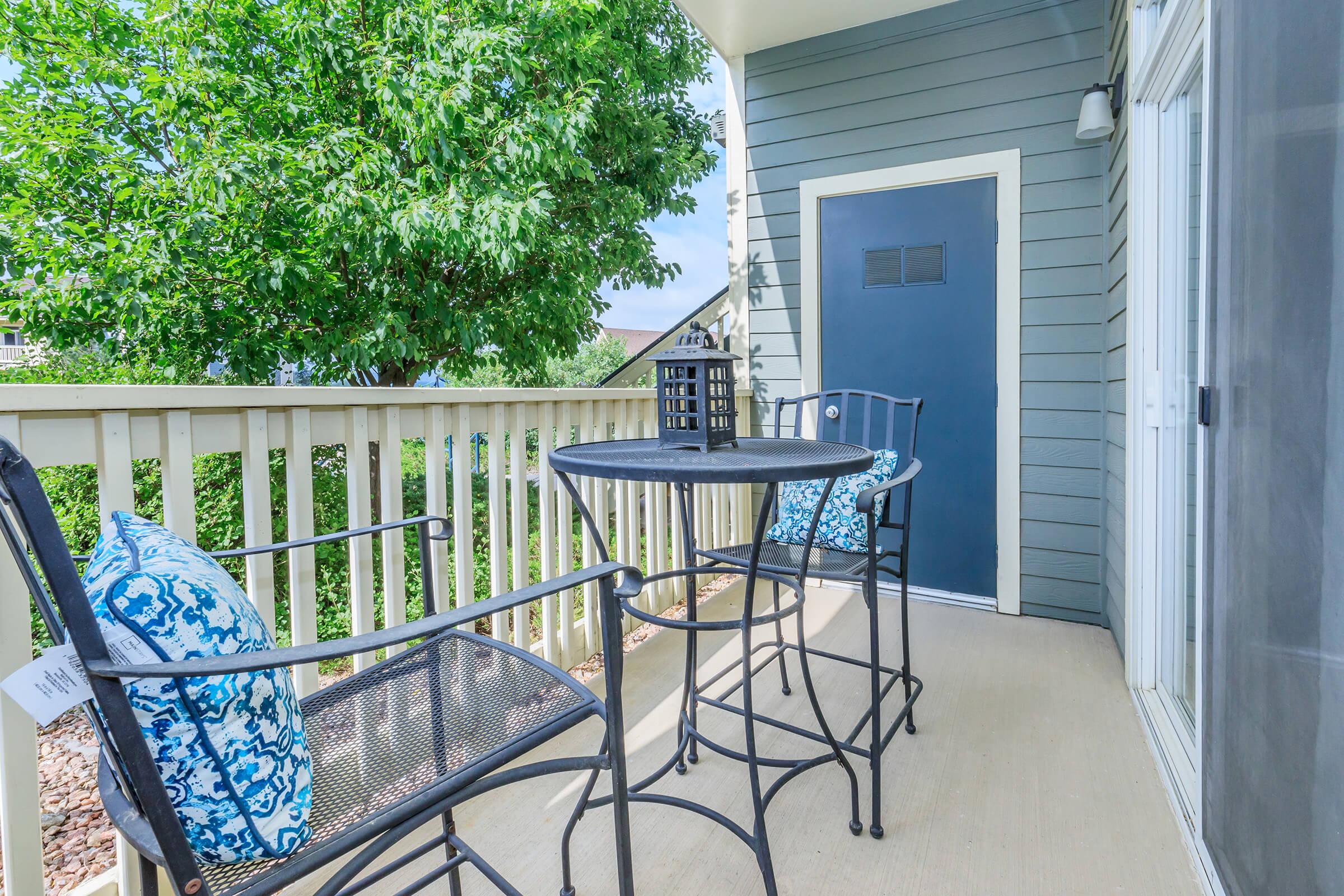 A cozy balcony featuring two high chairs with decorative blue pillows and a small round table. The backdrop includes a lush green tree and a door leading inside. Natural light brightens the space, creating a welcoming atmosphere for relaxation or outdoor dining.