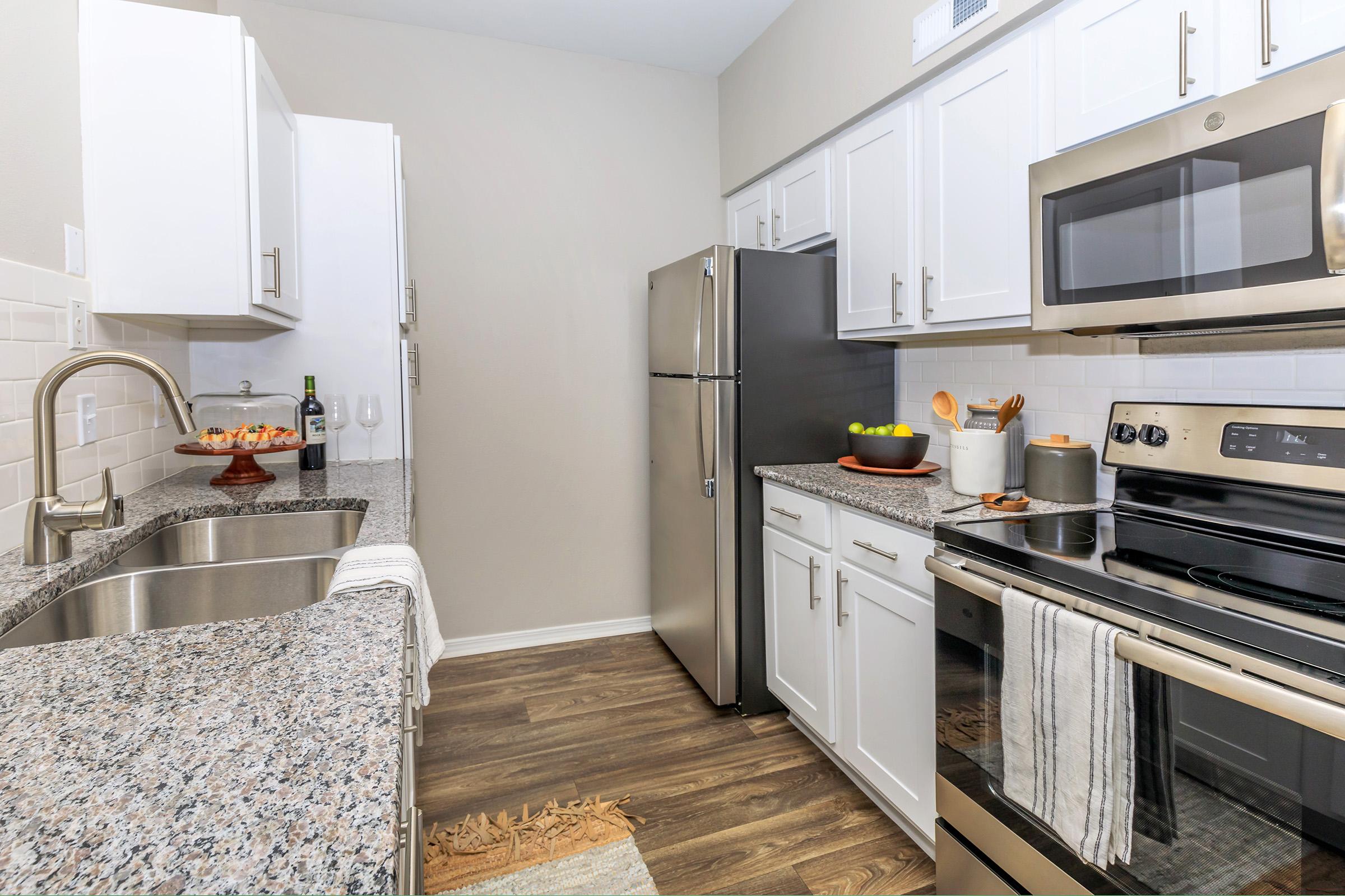 Modern kitchen featuring granite countertops, stainless steel appliances, and white cabinetry. The space includes a double sink, an electric stove, and a refrigerator. A decorative bowl with fruit sits on the counter, while a tray with wine glasses is visible. The flooring is a warm wood finish.