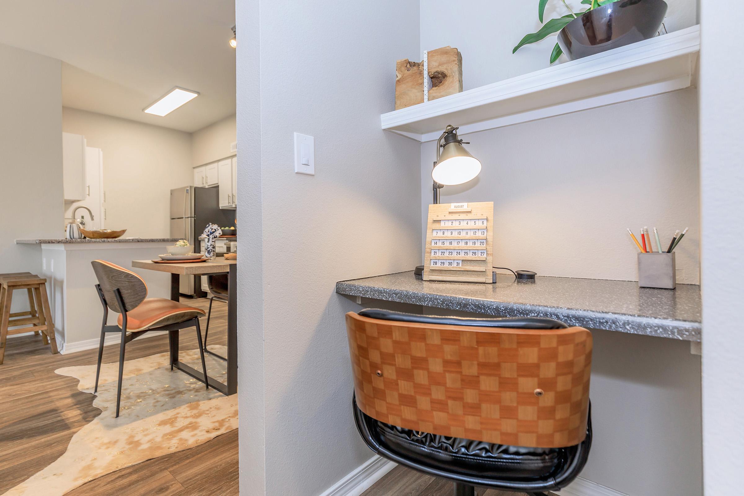 A small workspace featuring a desk with a lamp, a vintage-style calculator, and decorative items. The chair has a woven design, and there's a cozy dining area in the background with a table, chairs, and modern kitchen cabinets. Warm wooden flooring adds to the inviting atmosphere.