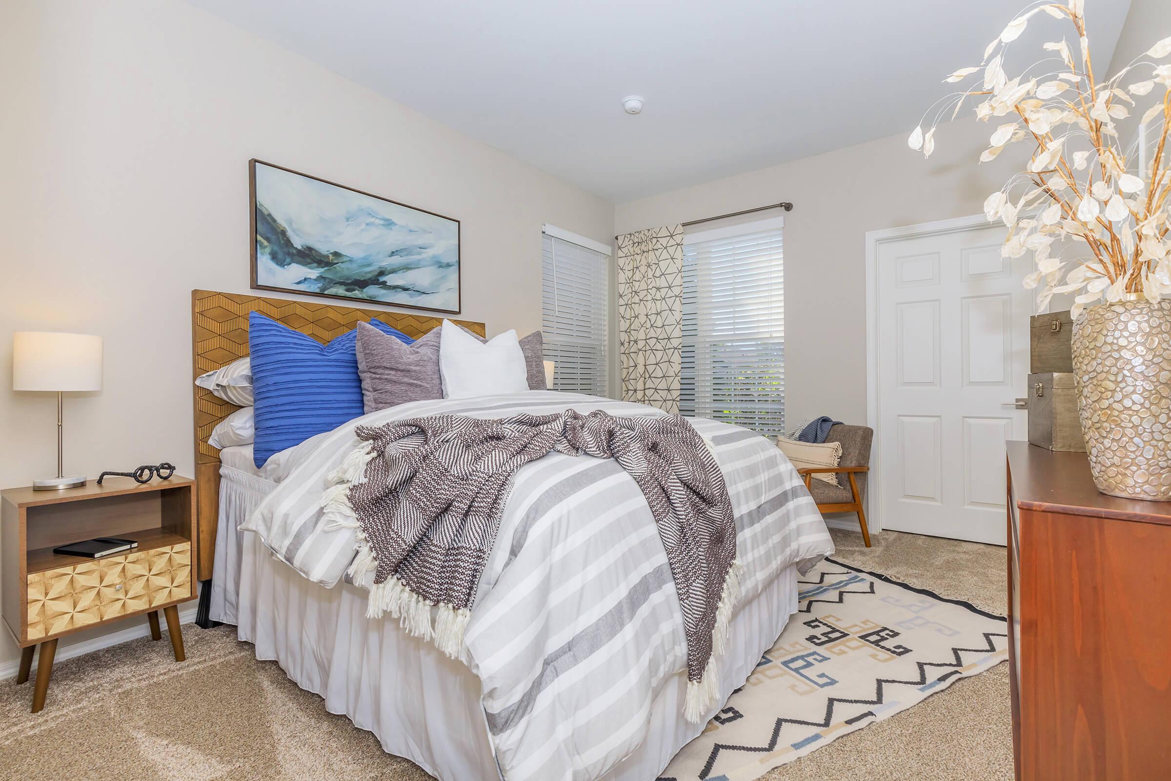 A cozy bedroom featuring a large bed with an array of blue and white pillows. The bed is dressed with a gray and white striped blanket. Next to it, a small nightstand with a lamp and glasses. A patterned area rug covers the floor, and a decorative plant adds a touch of warmth. Natural light enters through window treatments.