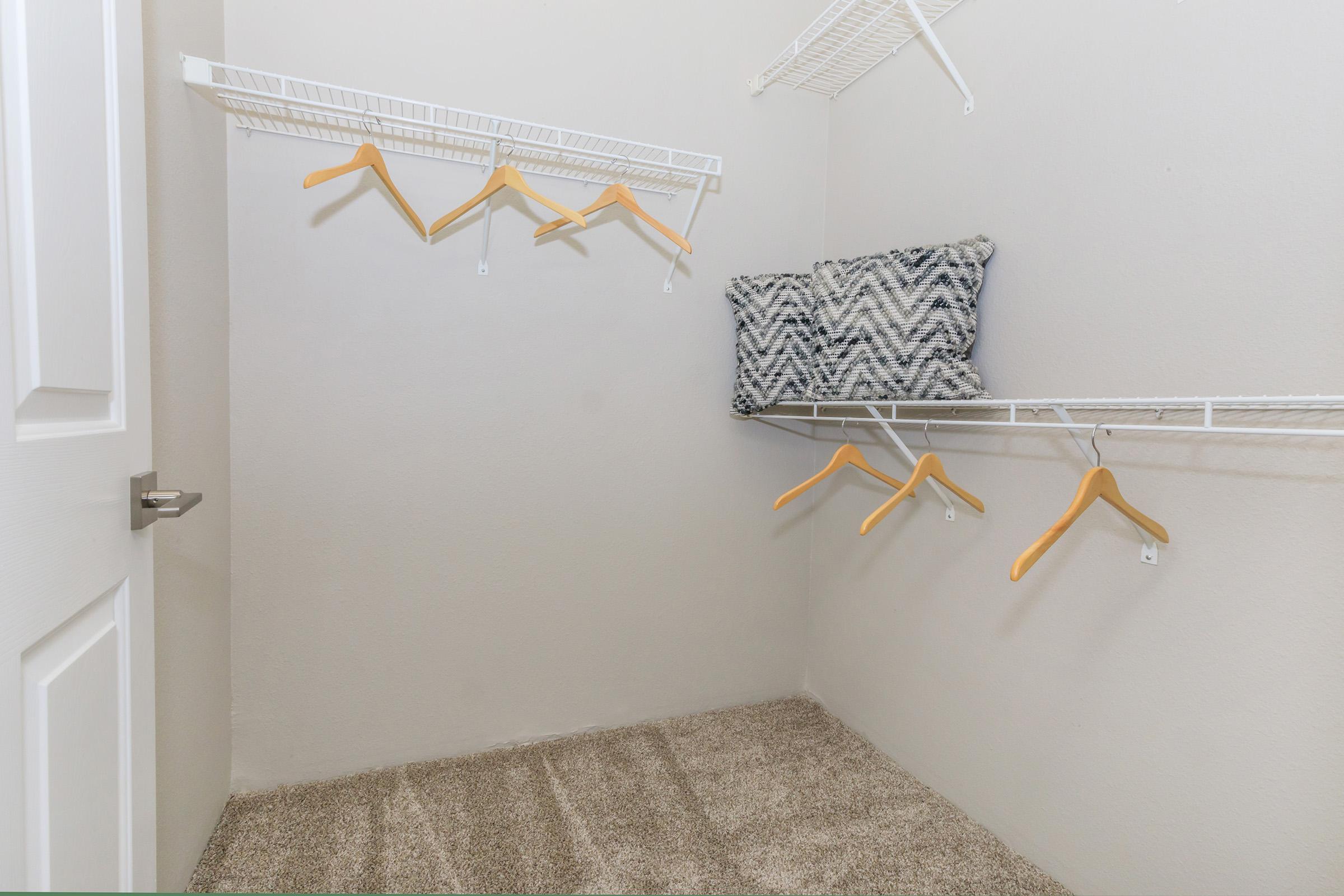 Empty closet with beige walls and carpet flooring. Features wire shelving with several wooden hangers and a patterned decorative pillow resting on the top shelf. The closet has a white door partially visible on the left.