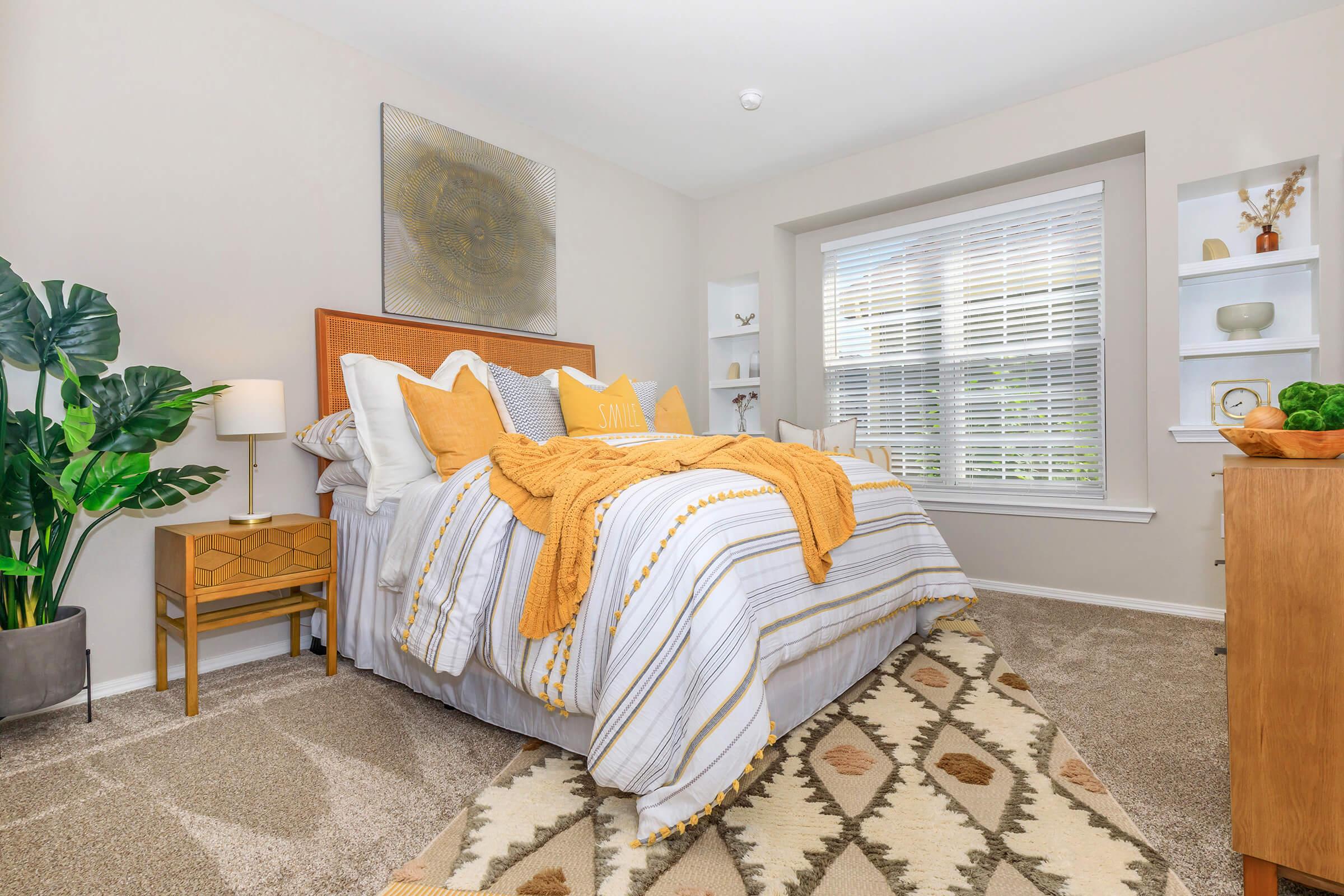 Bright and inviting bedroom featuring a neatly made bed with yellow and white bedding. A cozy textured blanket is draped over the bed. A green potted plant adds a touch of nature, while a bedside table holds a lamp. Natural light floods through the window, enhancing the warm aesthetic of the room.