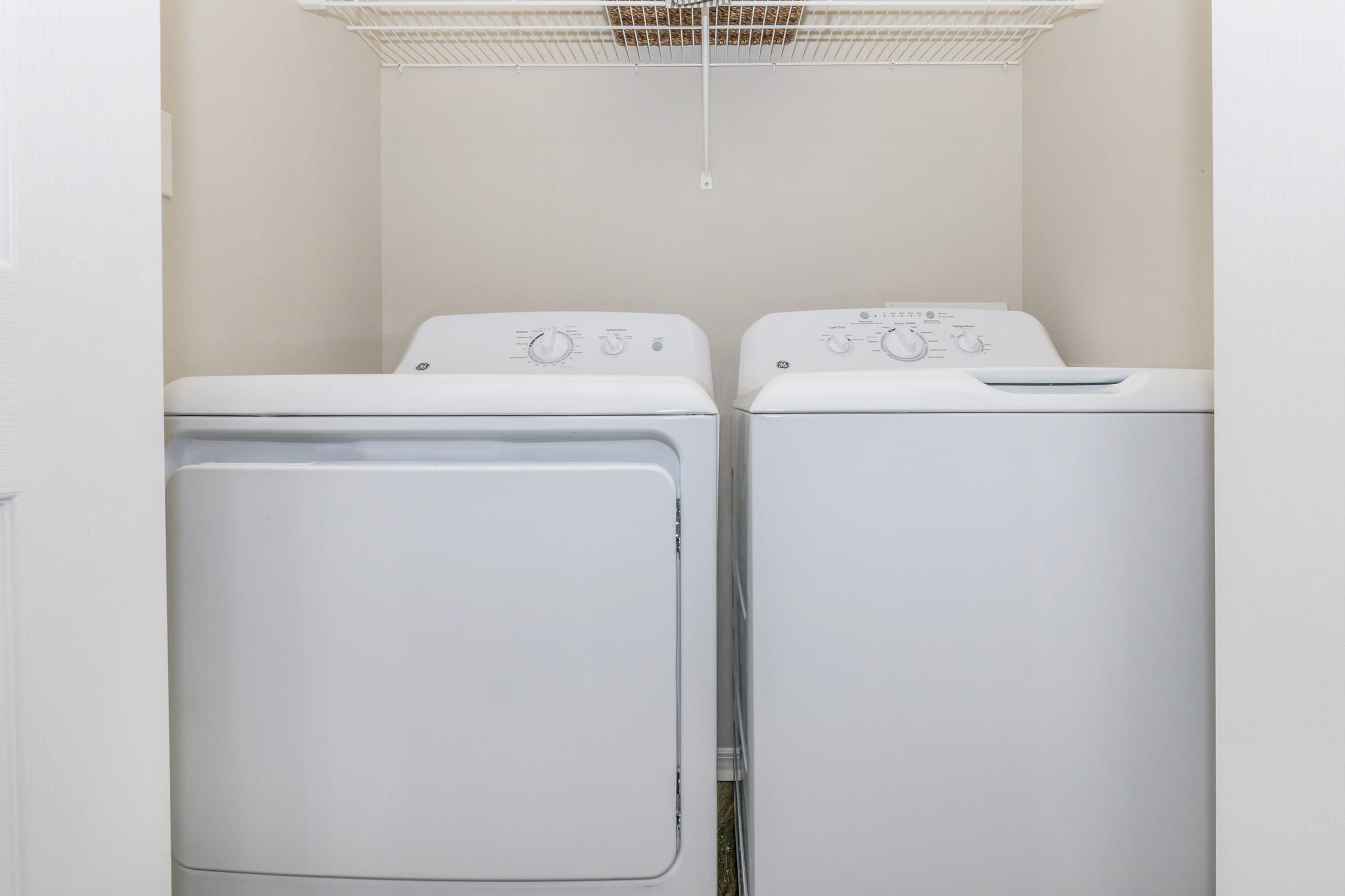 A laundry room featuring a white washing machine and a white dryer side by side. The appliances are placed in a small space with a shelf above for storage. The walls are light-colored, creating a clean and organized appearance.