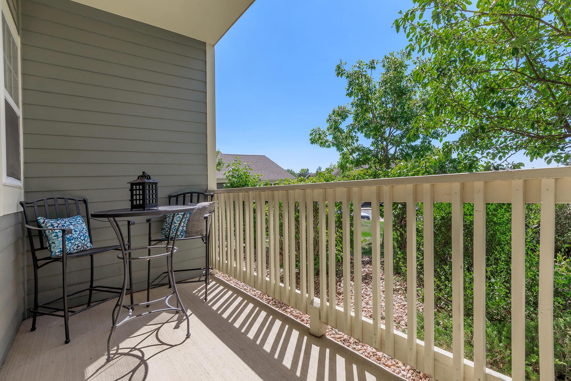 A small balcony featuring a round table and two high chairs. A black lantern is placed on the table, and blue paisley-patterned cushions are on the chairs. Sunlight casts shadows on the floor, and vibrant greenery is visible in the background against a clear blue sky.