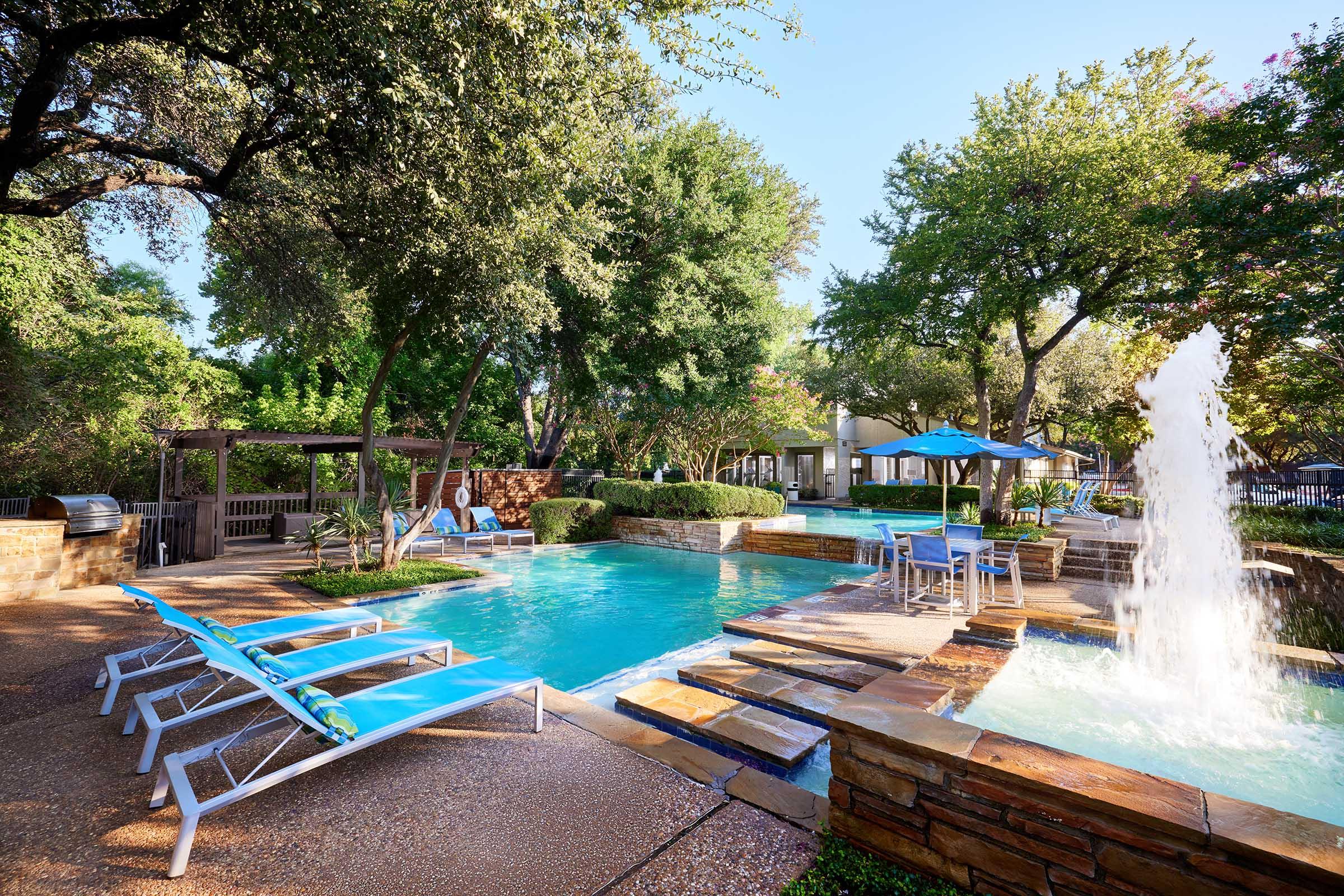 A serene outdoor pool area featuring lounge chairs and a gentle fountain surrounded by lush greenery and trees. The setting includes a well-maintained landscape with stone accents, creating a relaxing atmosphere perfect for leisure and enjoyment.
