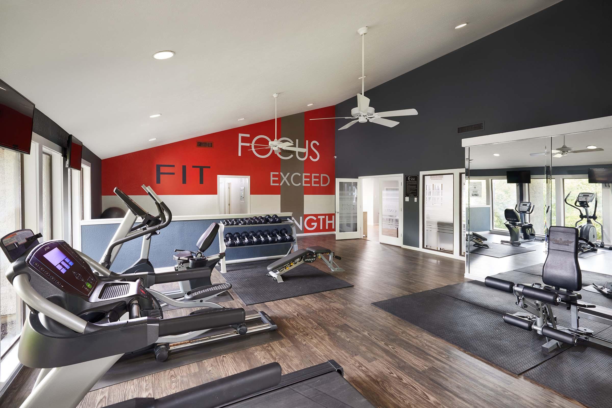 A modern gym interior featuring exercise equipment including treadmills, stationary bikes, and free weights. The walls are decorated with motivational phrases like "FOCUS," "FIT," "EXCEED," and "STRENGTH." Large windows allow natural light into the space, creating an inviting workout environment.