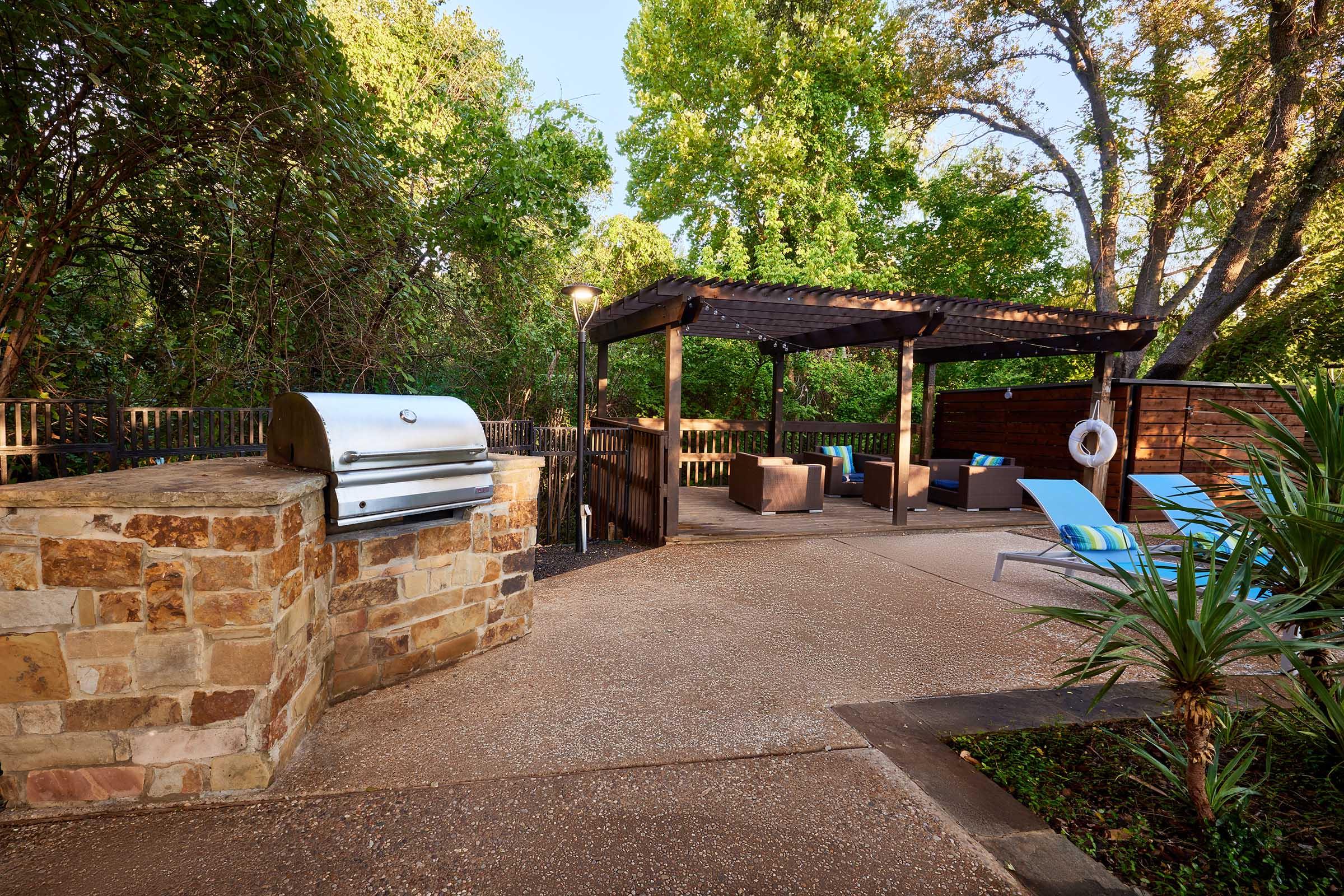 A serene outdoor area featuring a stone barbecue grill, a shaded pergola with seating, and poolside chairs. Lush greenery surrounds the space, creating a peaceful atmosphere, perfect for relaxation or entertaining. The scene invites outdoor enjoyment in a well-maintained setting.