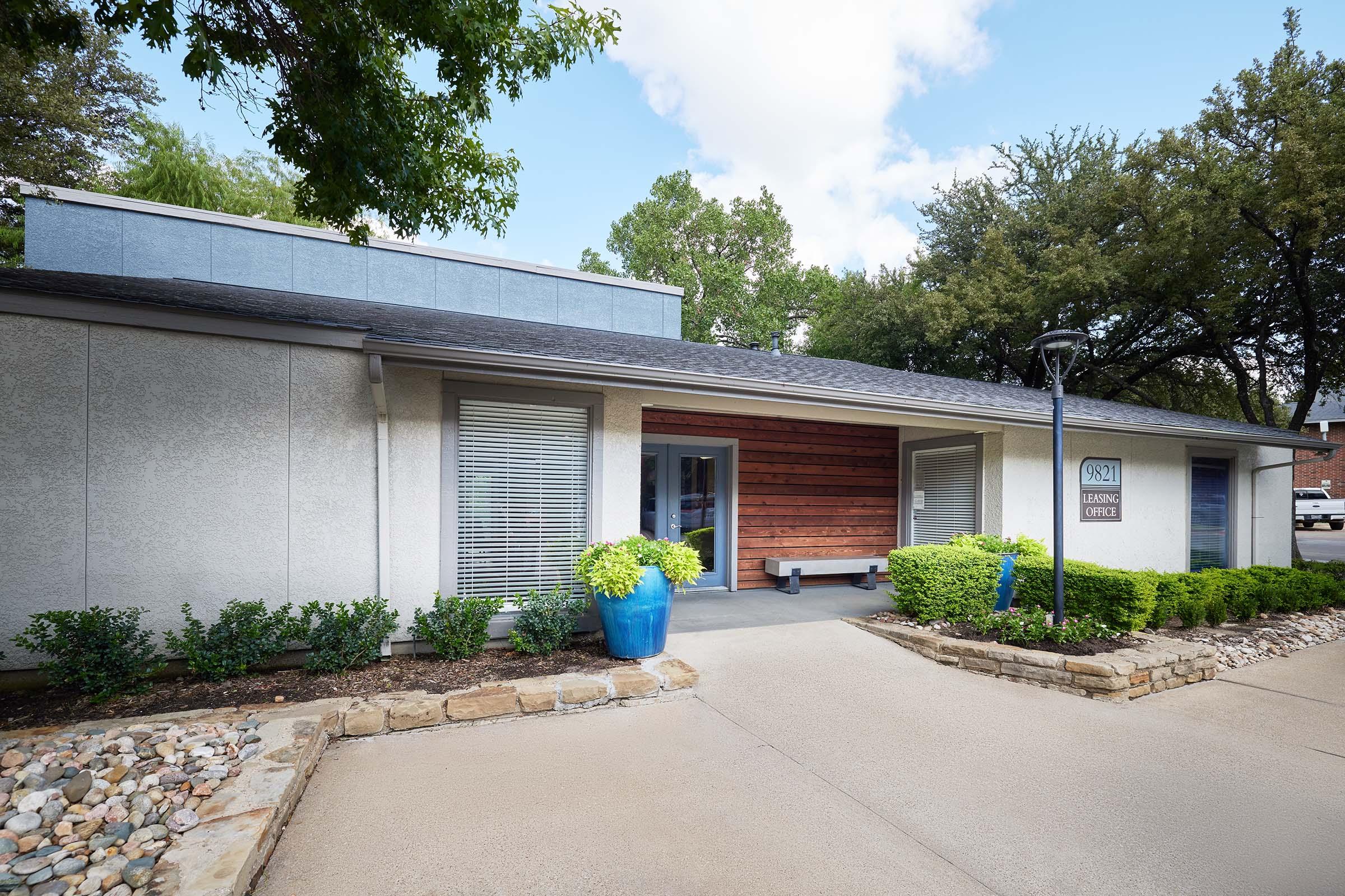 Exterior view of a modern leasing office with a mix of gray and wooden paneling, large windows with white blinds, and blue planters. The entrance features a wooden accent wall and a concrete path leading up to the door, surrounded by well-maintained greenery and stones. Bright sunlight illuminates the scene under a partly cloudy sky.
