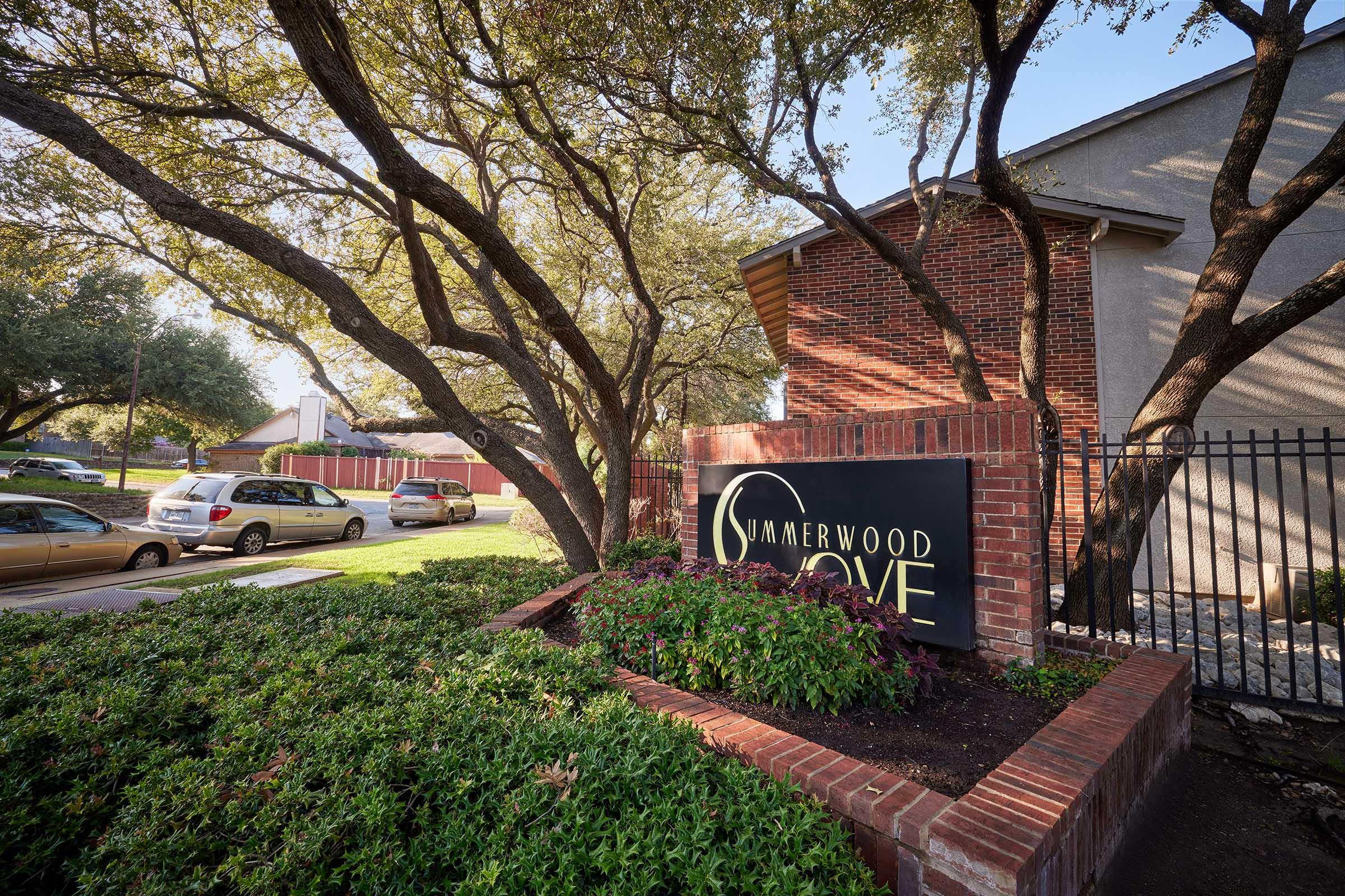 A brick sign reading "Summerwood Cove" is surrounded by lush greenery and decorative plants. Trees provide shade in the foreground, and parked cars are visible along a paved area. The setting appears to be a residential community or complex.