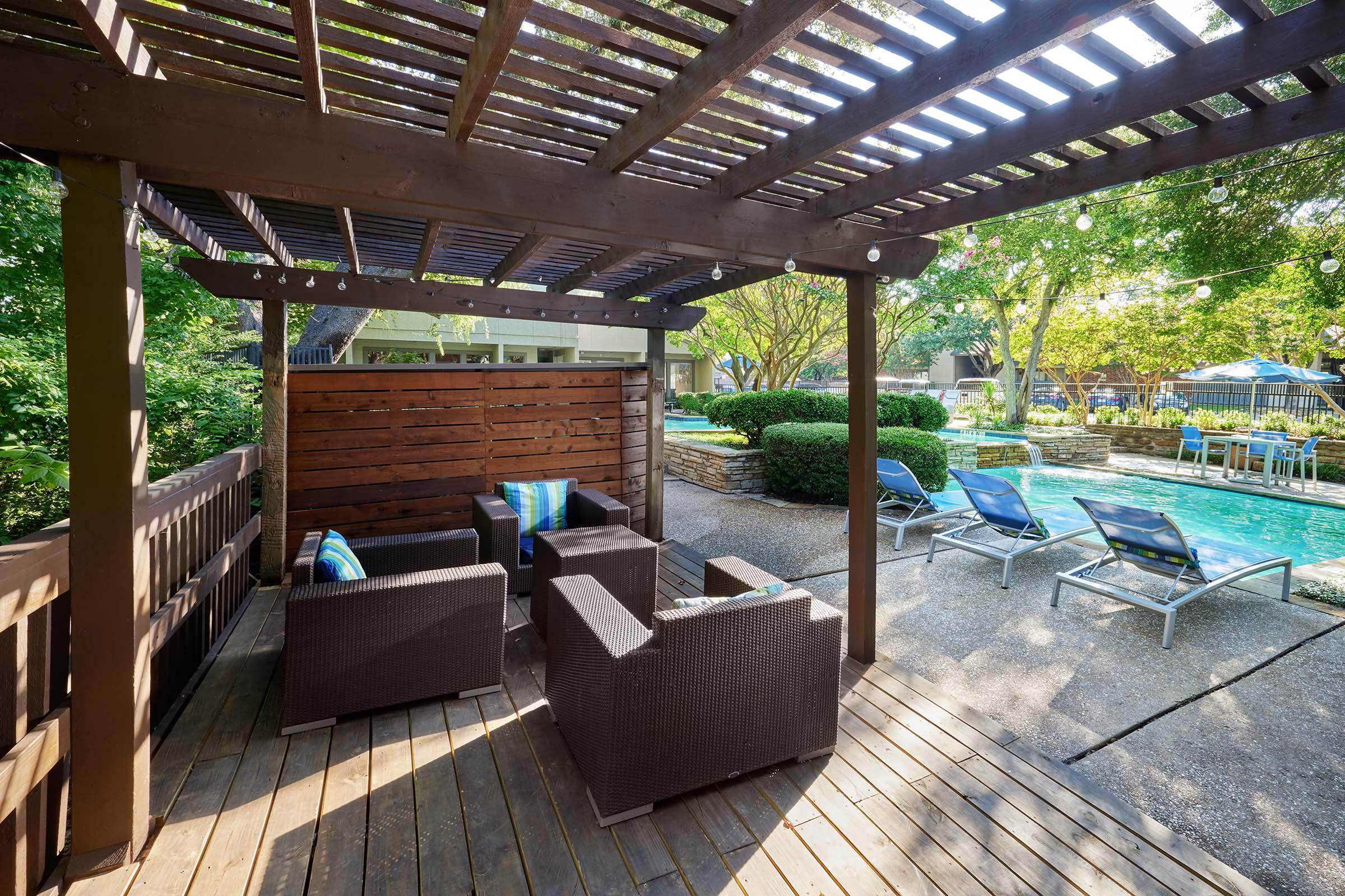 A cozy outdoor seating area under a wooden pergola, featuring dark wicker chairs and blue cushions. In the background, a sparkling swimming pool is surrounded by lounge chairs and lush greenery, creating a relaxing and inviting atmosphere for leisure.