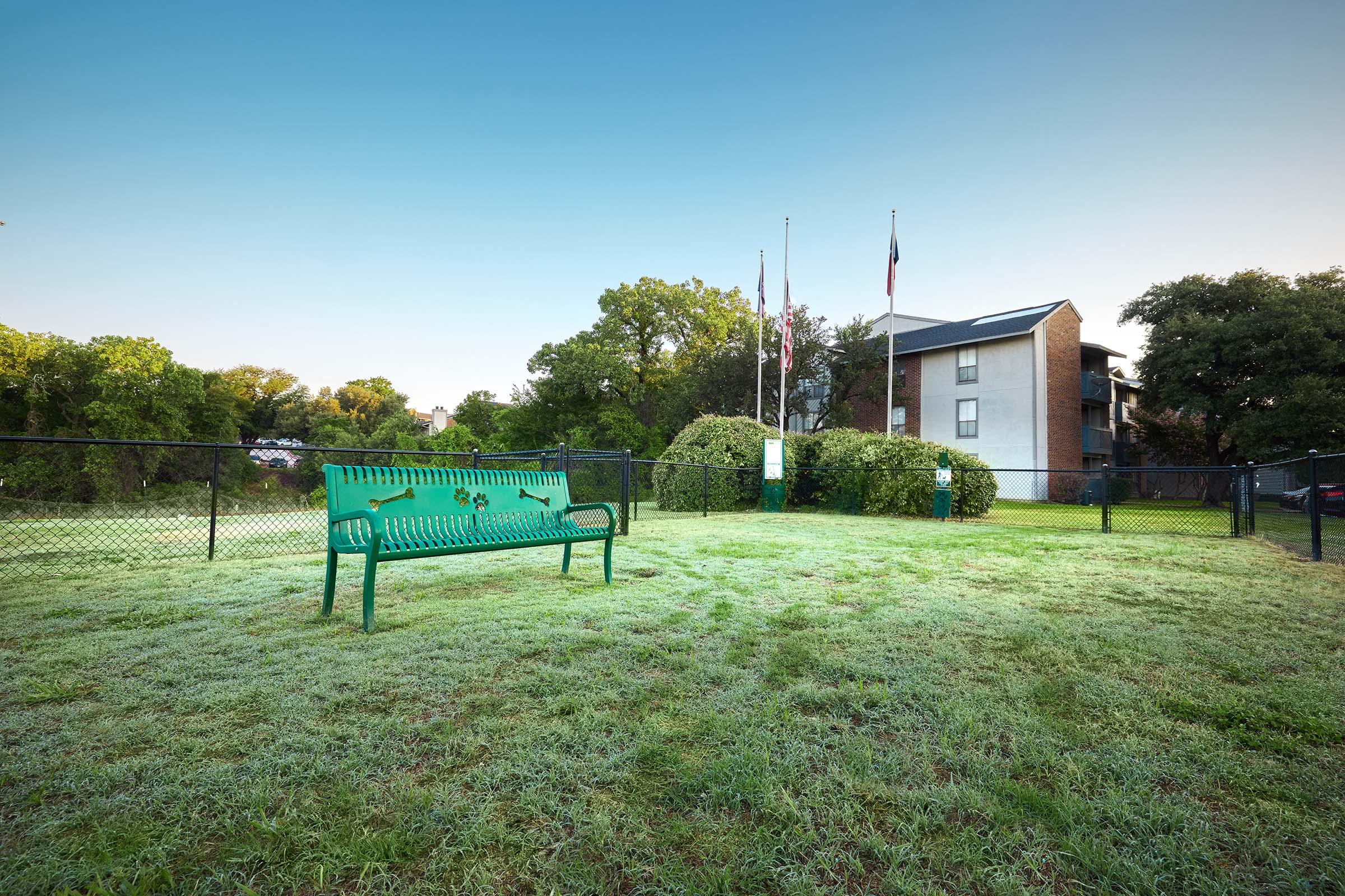 A green park bench sits on a grassy area surrounded by a black fence, with flags at half-mast in the background. A multi-story apartment building is visible behind the bench, and the setting appears tranquil with trees and an open sky.