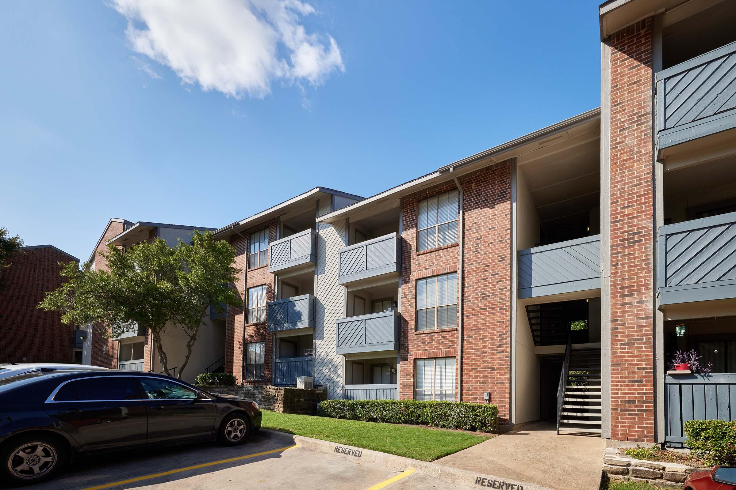 Brick apartment building with multiple balconies and stairs, surrounded by greenery. A parked car is in the foreground, and a clear blue sky with a few clouds is in the background. The setting appears to be a residential area.