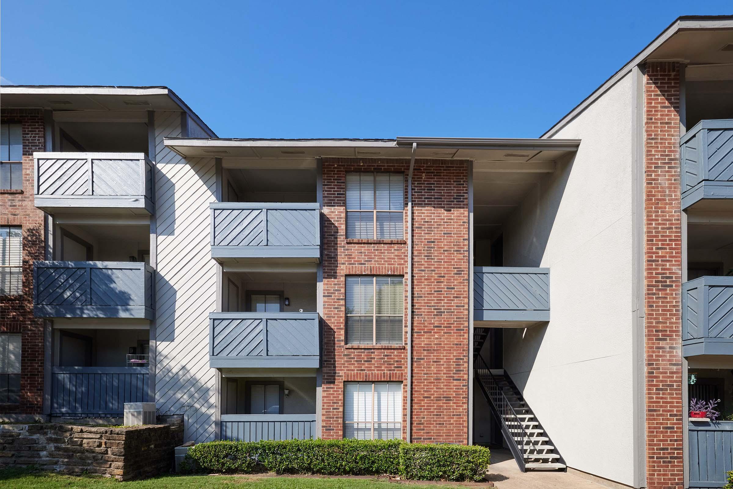 Three-story apartment building with a brick facade and gray balconies. Front entrance features a stairway leading to upper levels. Lush green landscaping in front under a clear blue sky.