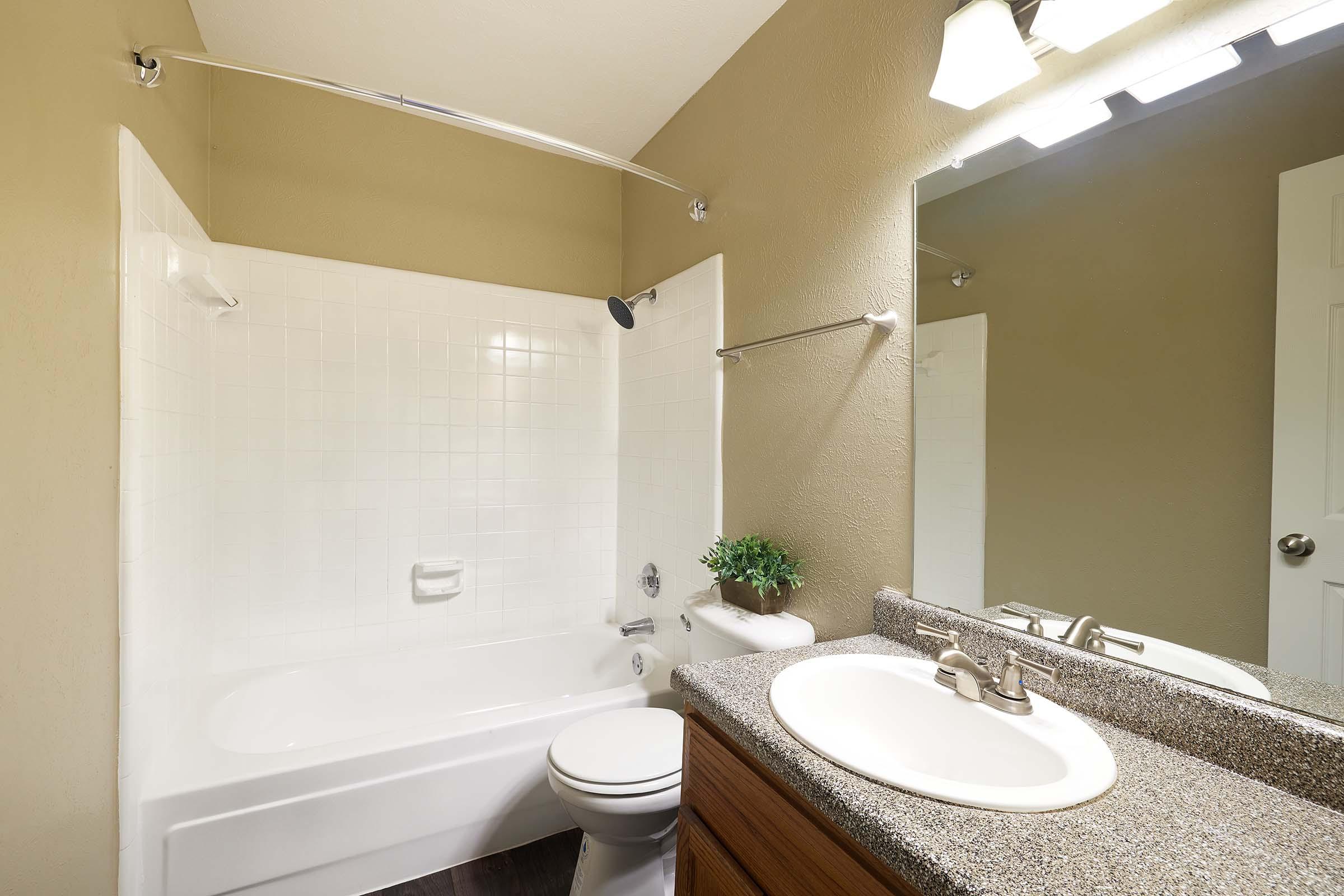 A spacious bathroom featuring a bathtub with a white tile surround, a light-colored countertop with a sink, and a large mirror above the sink. A small potted plant adds decoration, while the walls are painted a warm shade. Modern fixtures and a shower rod are also visible.