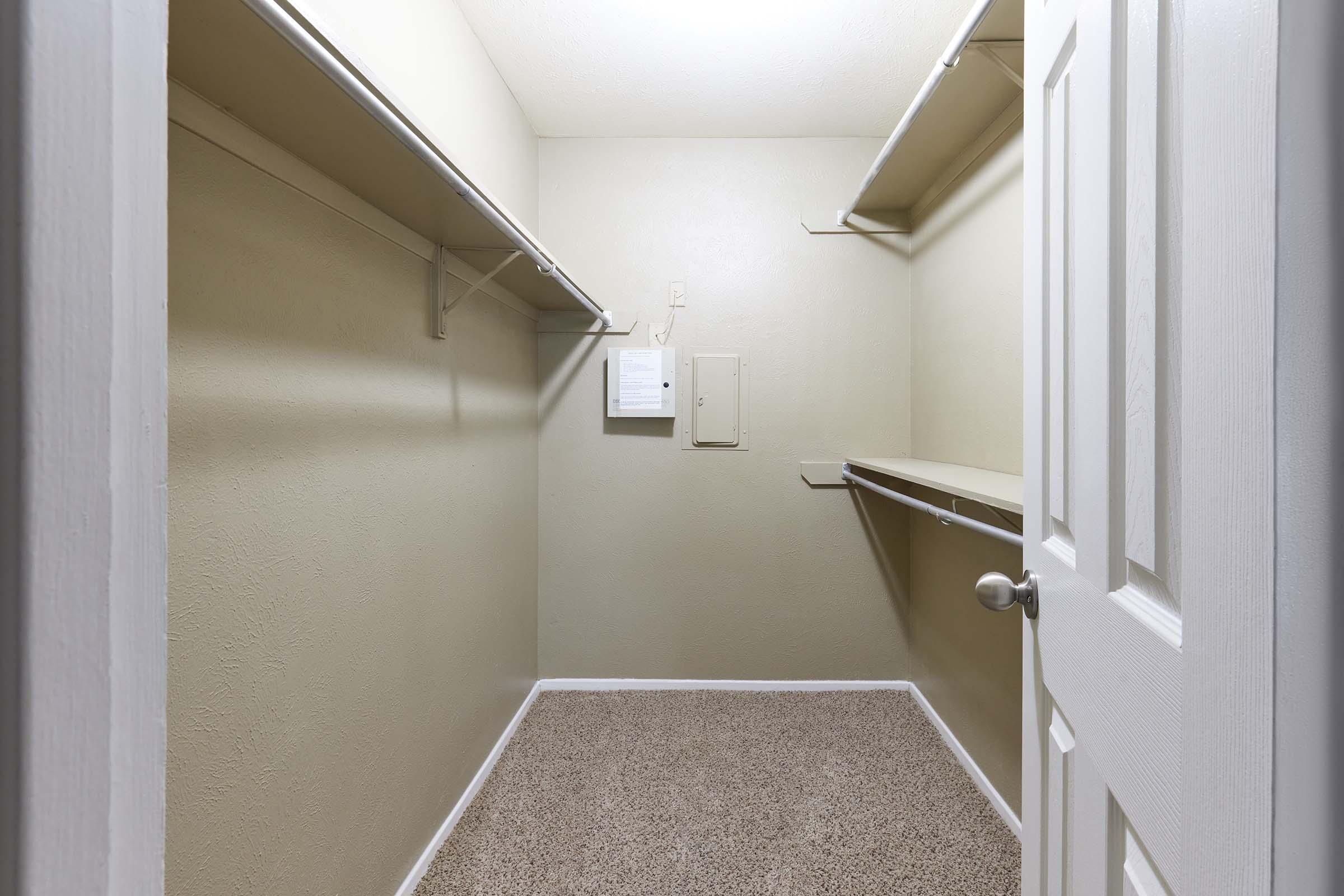 Empty closet with beige walls and carpeted floor. It features white shelving on one side and a small electrical panel on the opposite wall. The door is partially open, showing the interior space that lacks storage items or decor.