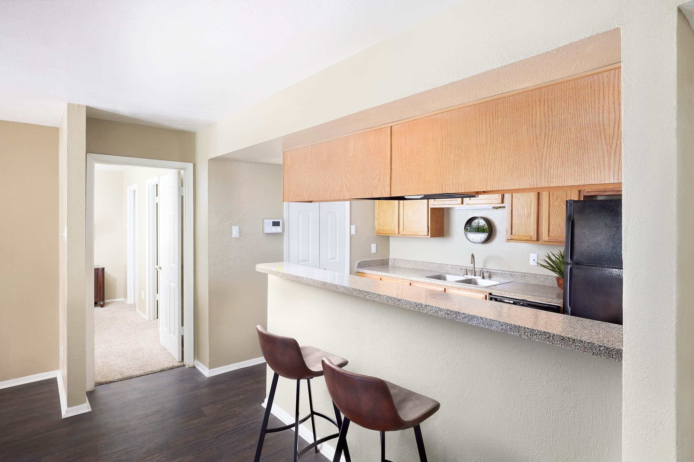 A modern kitchen with wooden cabinets and a granite countertop, featuring a sink and appliances. There are two brown barstools at the edge, overlooking a hallway leading to rooms with white doors. The walls are painted in a neutral color, creating a bright and inviting atmosphere.