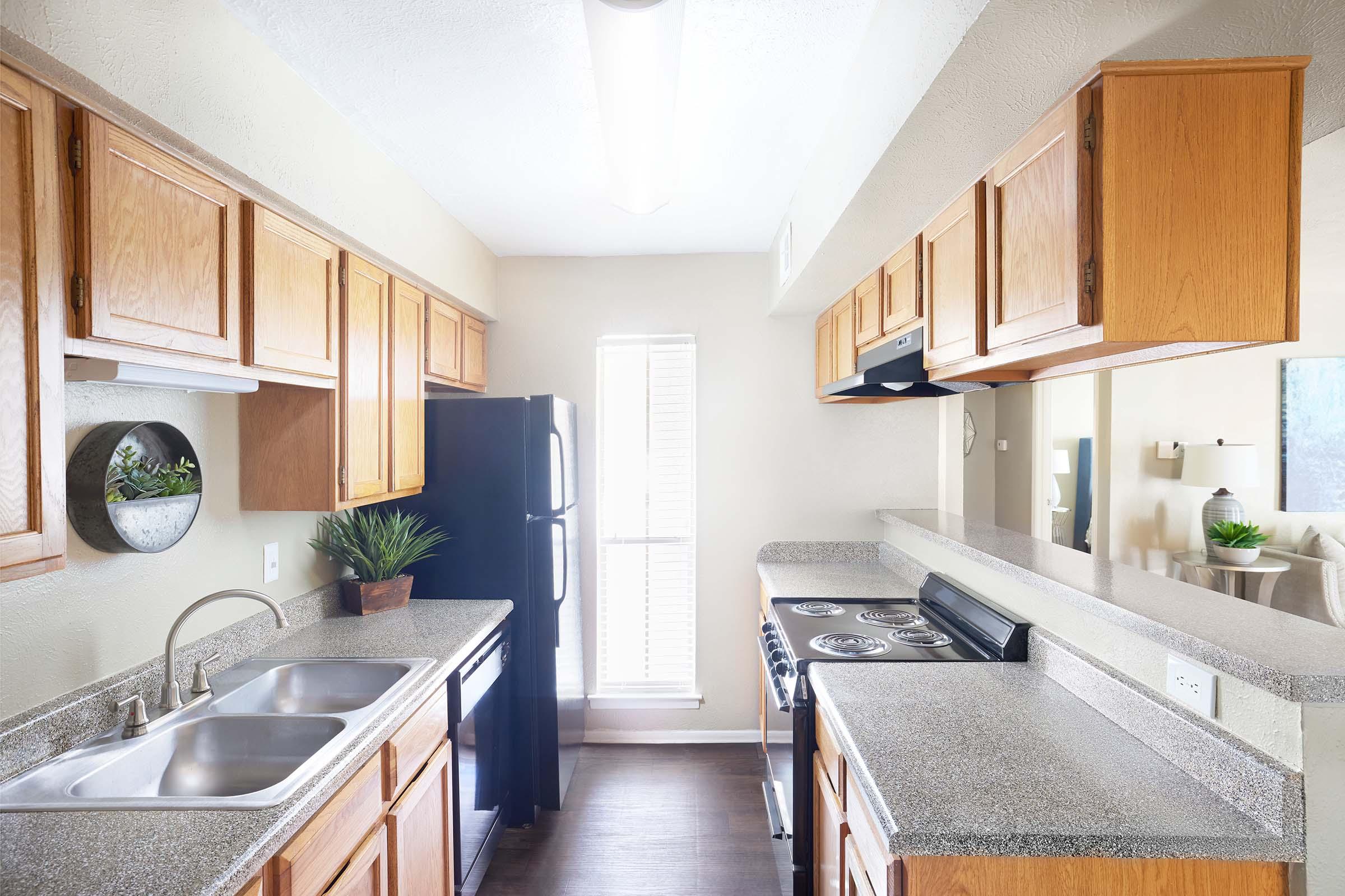 A modern kitchen featuring wooden cabinets, a double sink, and a black refrigerator. The countertops are grey and the space is well-lit with a window in the background. There are plants on the counter and a mirror reflecting part of the room, creating a welcoming atmosphere.