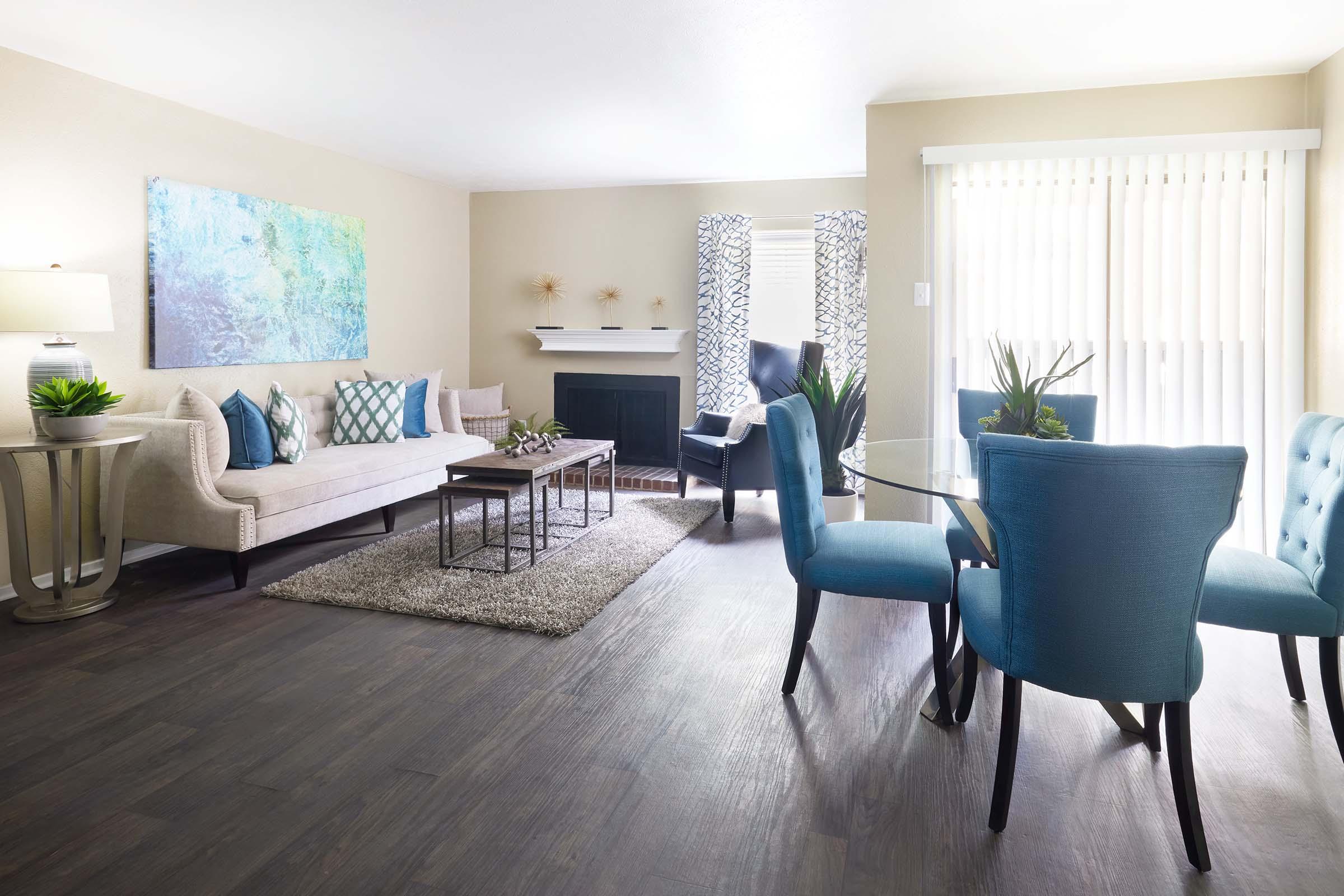 A bright, modern living room featuring a beige sofa with decorative pillows, a glass coffee table, and a cozy area rug. There are two stylish chairs and a round dining table with blue upholstered chairs. A wall art piece adds color, while large windows provide natural light.