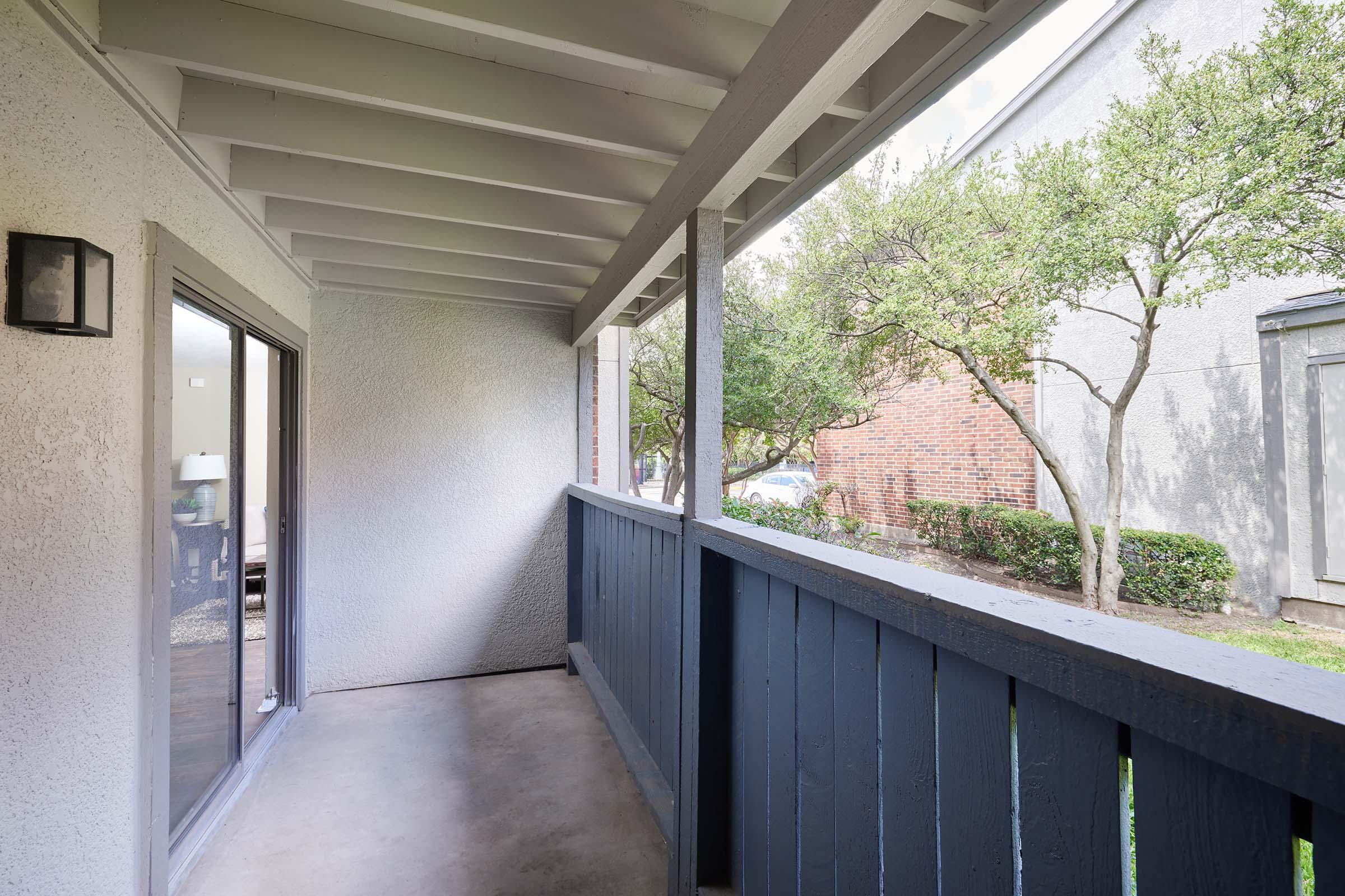 A small covered balcony with a gray railing, featuring a concrete floor and white walls. Natural light filters in from sliding glass doors, and green trees and shrubs are visible outside, creating a serene outdoor view.