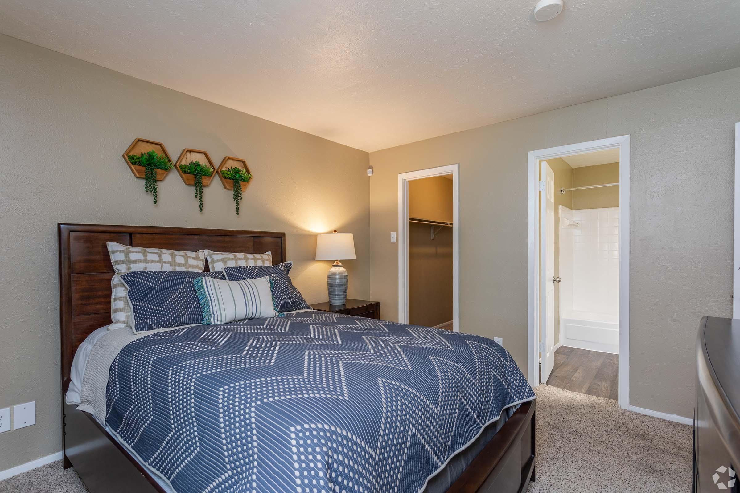 A cozy bedroom featuring a wooden bed with a blue and white patterned comforter. To the left, there is a bedside lamp on a nightstand, with three decorative wall hangings above the bed. A doorway leads to a closet, and another door opens to a bathroom. The walls are painted in a soft, neutral color.