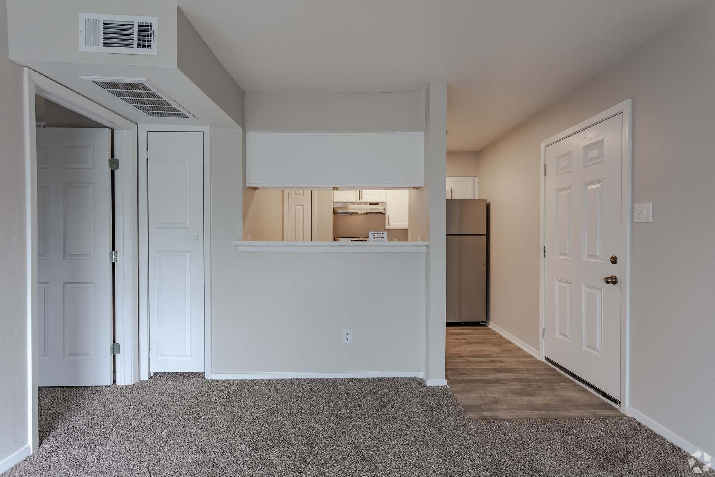 Interior view of an apartment showcasing an open layout. A doorway is visible on the left leading to a room, with a pass-through counter that opens into a kitchen area. The front door is on the right, and light-colored walls and carpeted flooring create a cozy atmosphere.