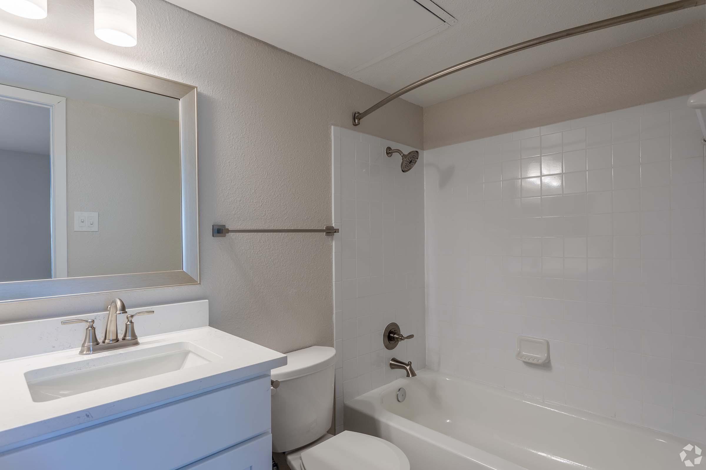 A modern bathroom featuring a white bathtub, shower with a glass screen, a toilet, and a sink with a mirror above. The walls are light-colored tiles, and there is a chrome showerhead and towel bar. Bright overhead lighting enhances the clean and spacious look.