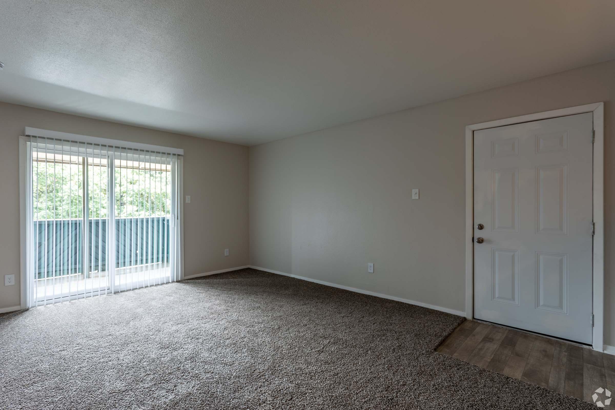 Empty living room with light beige walls and plush, brown carpeting. A large sliding glass door leads to an outdoor balcony area, brightening the space with natural light. The room features a neutral color palette, offering a blank canvas for furnishing and decorating. An entry door is visible on the right.