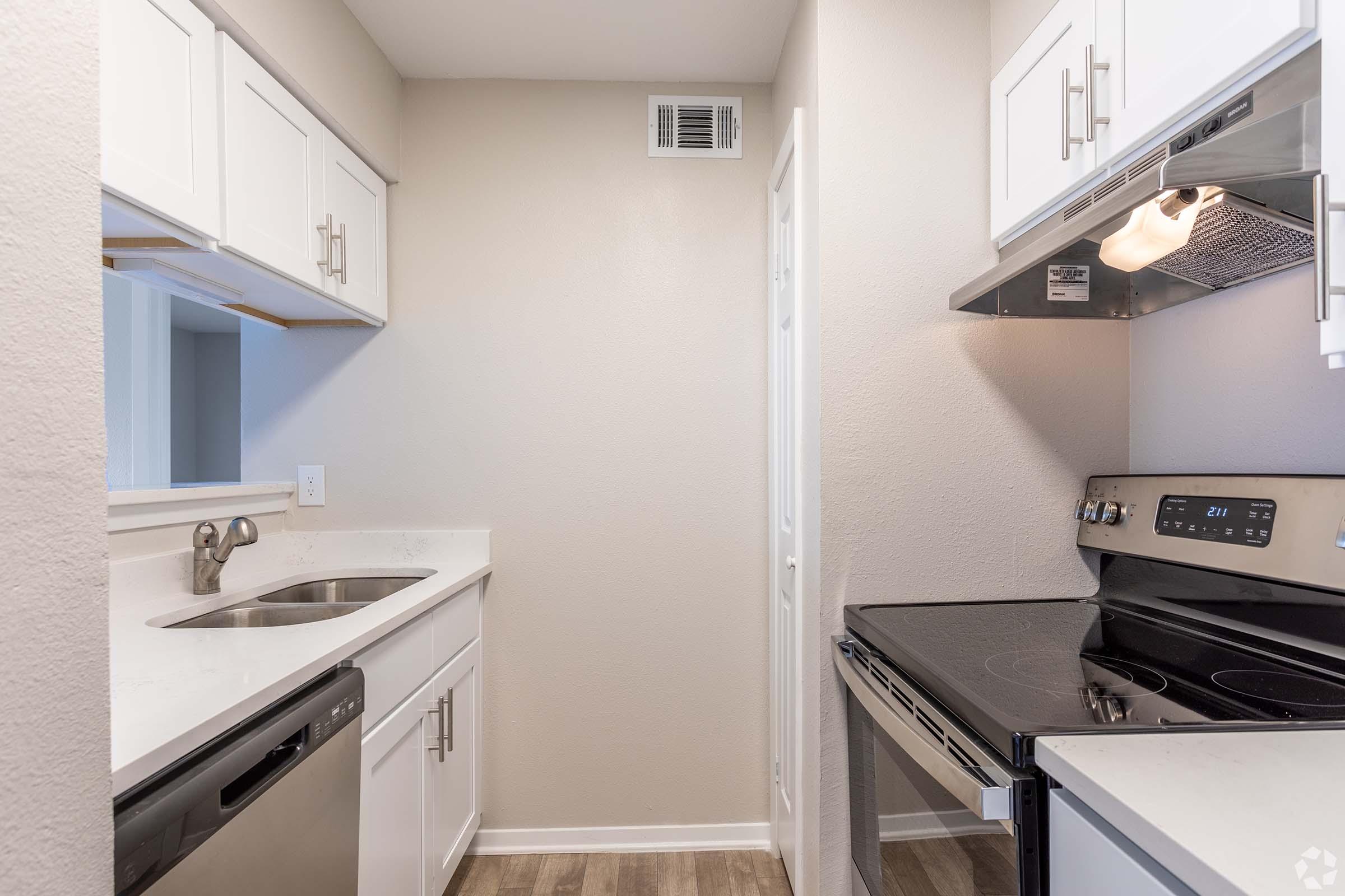 A modern kitchen featuring white cabinetry, a stainless steel sink, a black electric stove, and a dishwasher. The walls are painted in a light color, and there is a small window providing natural light. The flooring is a light wood laminate, giving the space a fresh and clean appearance.