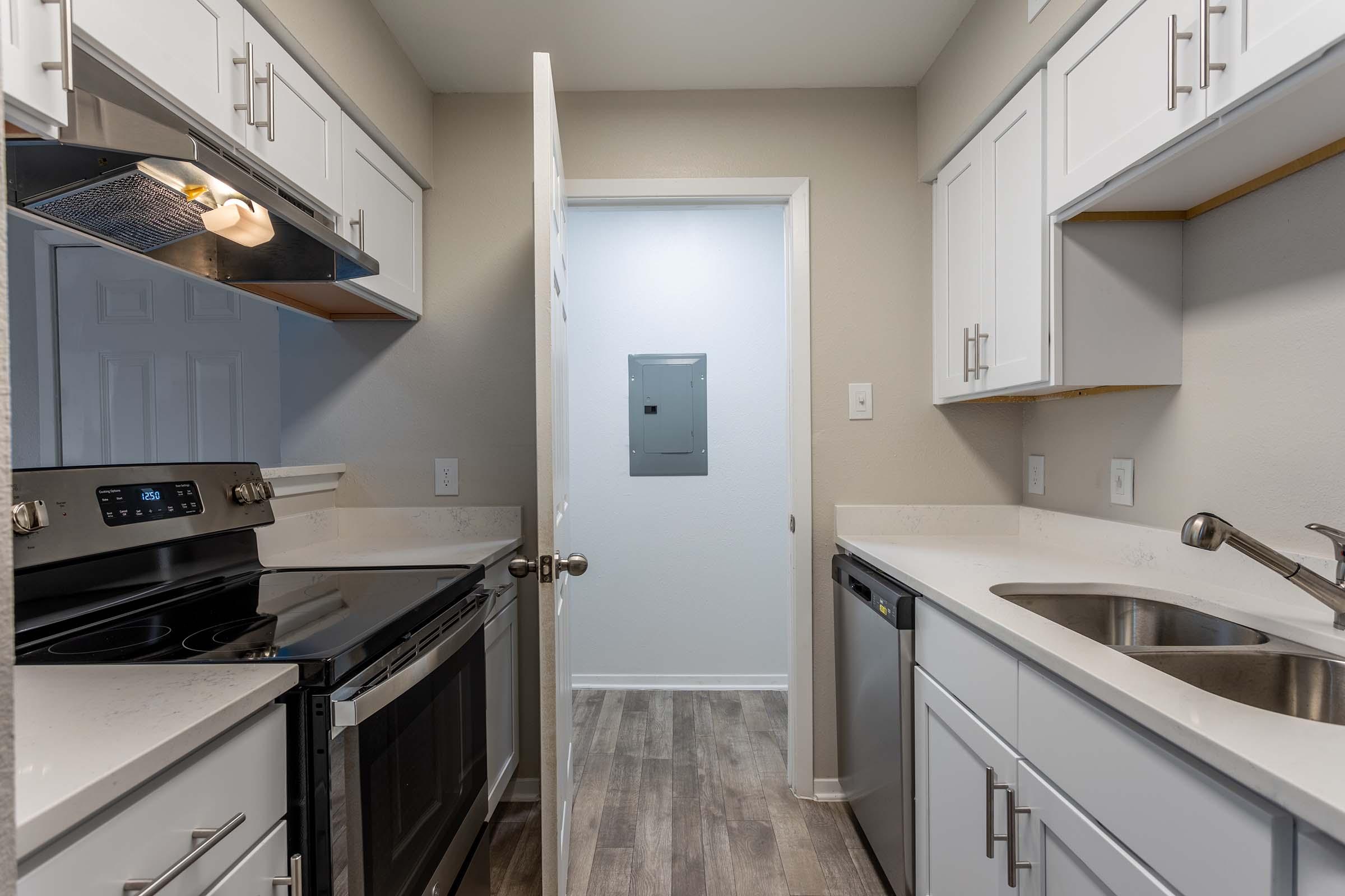 A modern kitchen featuring white cabinetry, stainless steel appliances including an oven and dishwasher, and a light-colored countertop. The space has a door leading to an adjacent room with an electrical panel, and warm wood-like flooring throughout.