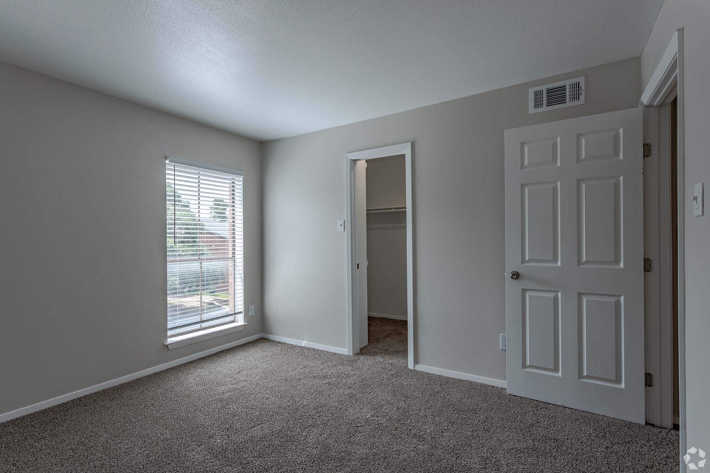 A light-colored room with beige carpet and a window featuring white blinds. There is a door leading to a closet on the right, and a neutral-colored wall opposite the window. The room has a bright, airy atmosphere with soft lighting.