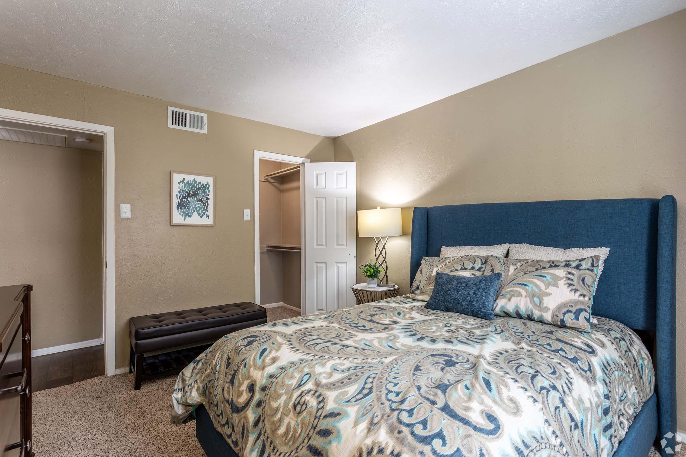 A cozy bedroom featuring a blue upholstered bed with patterned bedding, a decorative lamp beside it, and a dark bench at the foot of the bed. The room has light brown walls and a closet door visible on the left side, creating a warm and inviting atmosphere.