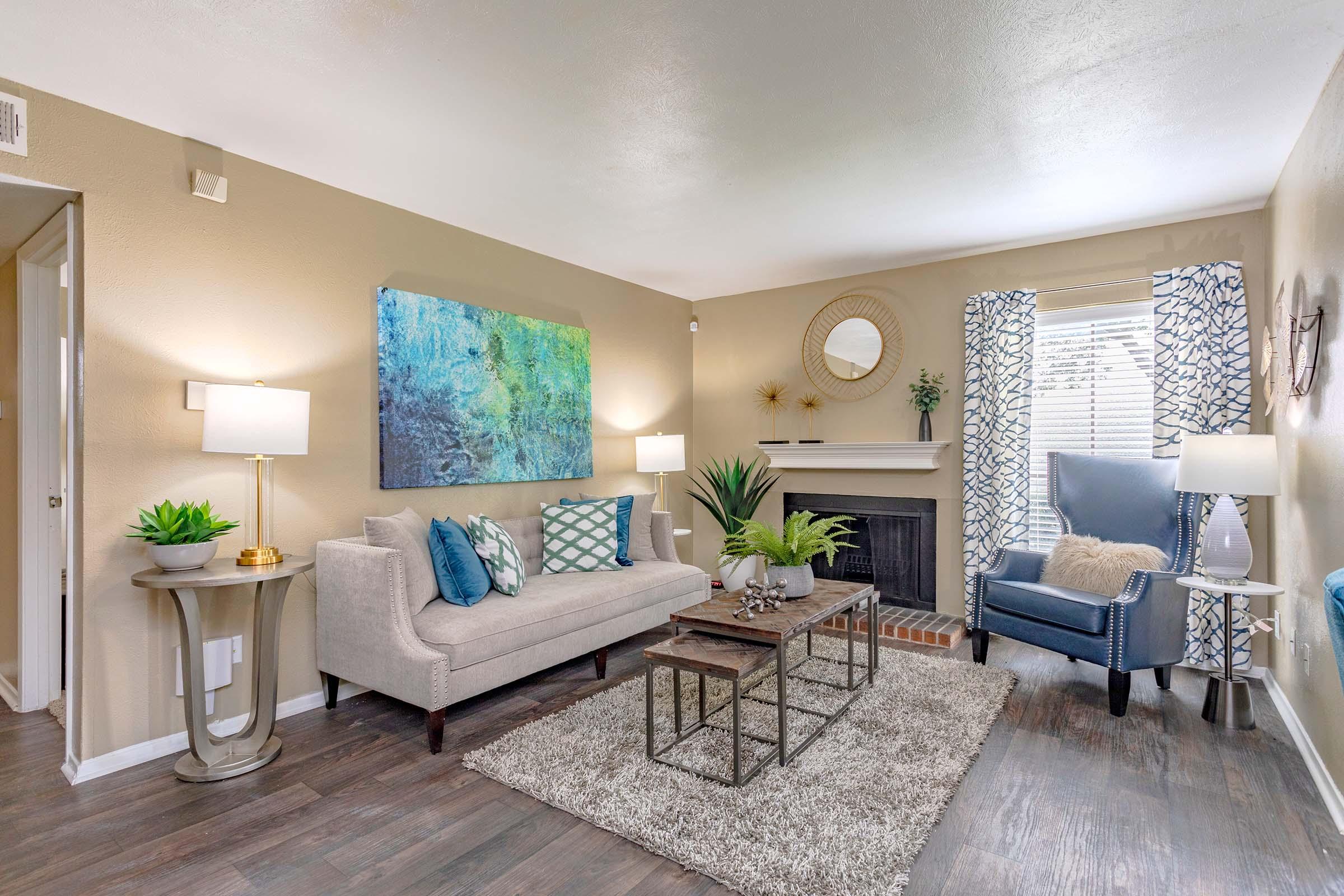 A cozy living room featuring a light gray sofa with decorative pillows, a coffee table with a metal frame, and a plush area rug. There are two lamps for lighting, a large abstract painting on the wall, and a blue accent chair to the side. Windows draped with patterned curtains allow natural light in, creating a welcoming atmosphere.